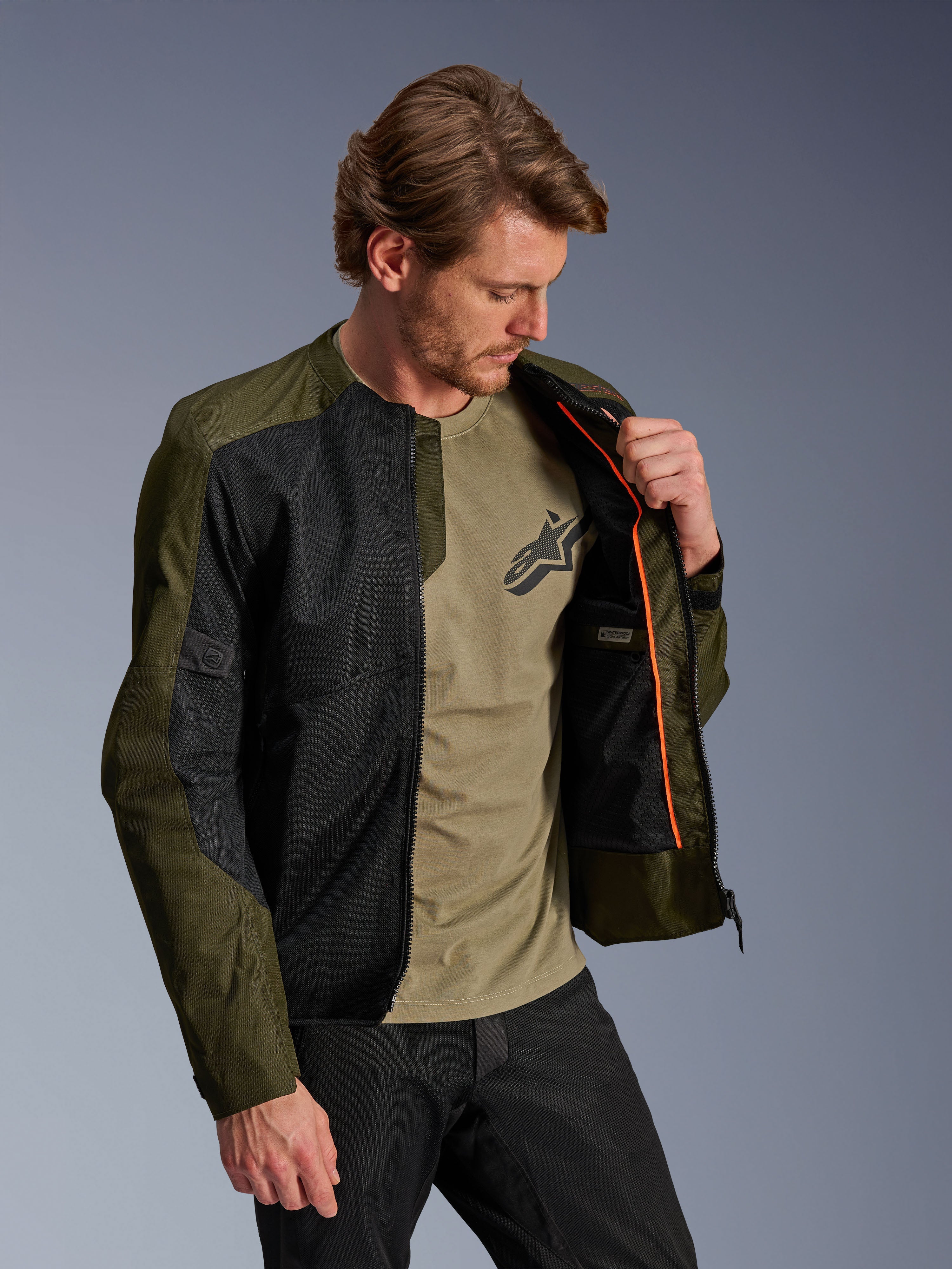 Casaco Alpinestars C-1 Air em caqui e preto, modelo masculino a mostrar o forro interior laranja e o compartimento impermeável, painéis de malha técnica para fluxo de ar, estilo urbano contemporâneo com t-shirt verde azeitona e calças pretas