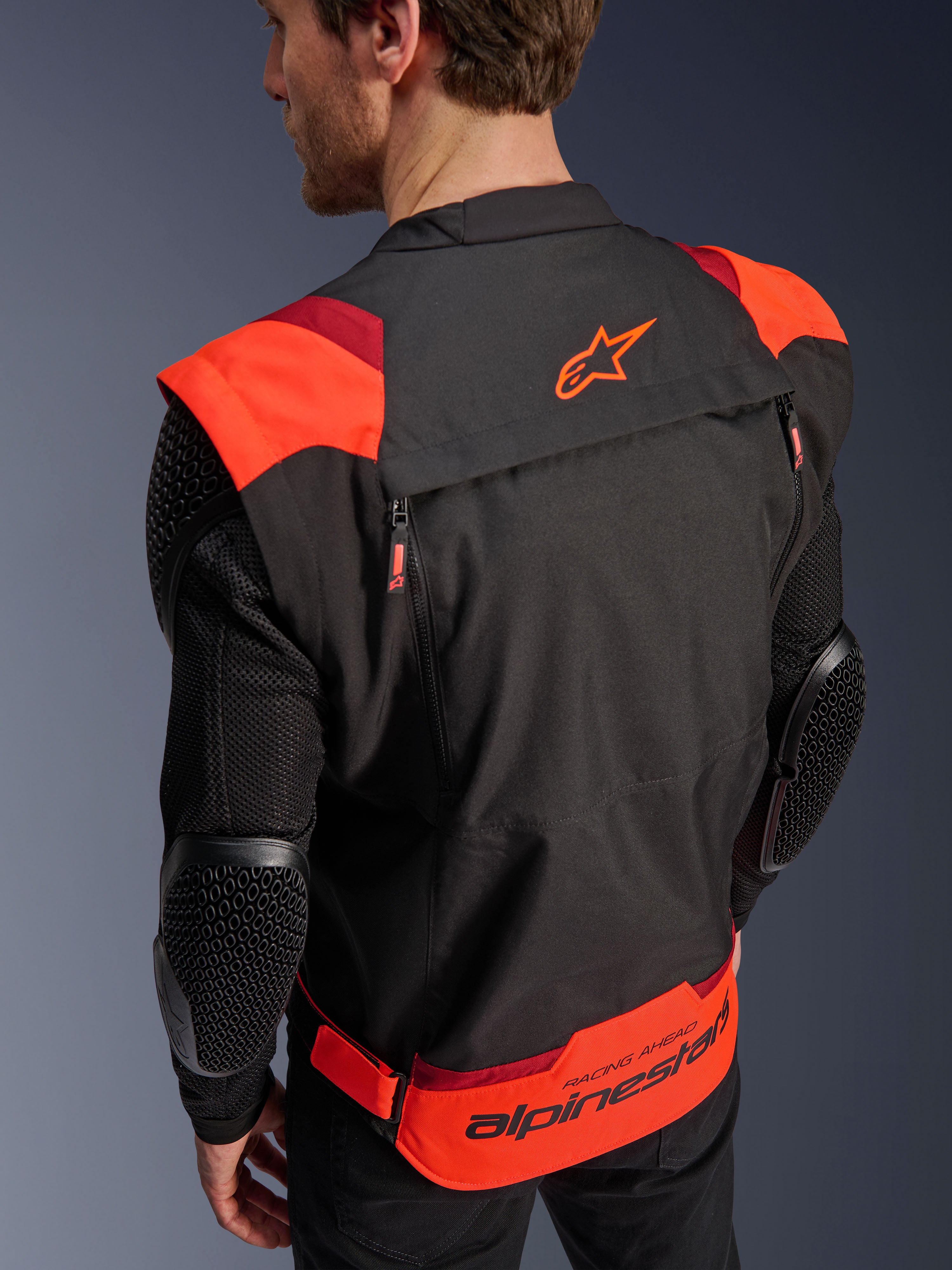 Casaco Alpinestars T-Stunt Air, preto, vermelho fluo e vermelho escuro, vista traseira num modelo, apresentando mangas em malha com protetores de cotovelo com textura em favo de mel, logótipo de estrela na parte superior das costas e marca 'RACING AHEAD' na zona lombar, design modular de corte desportivo para motociclismo