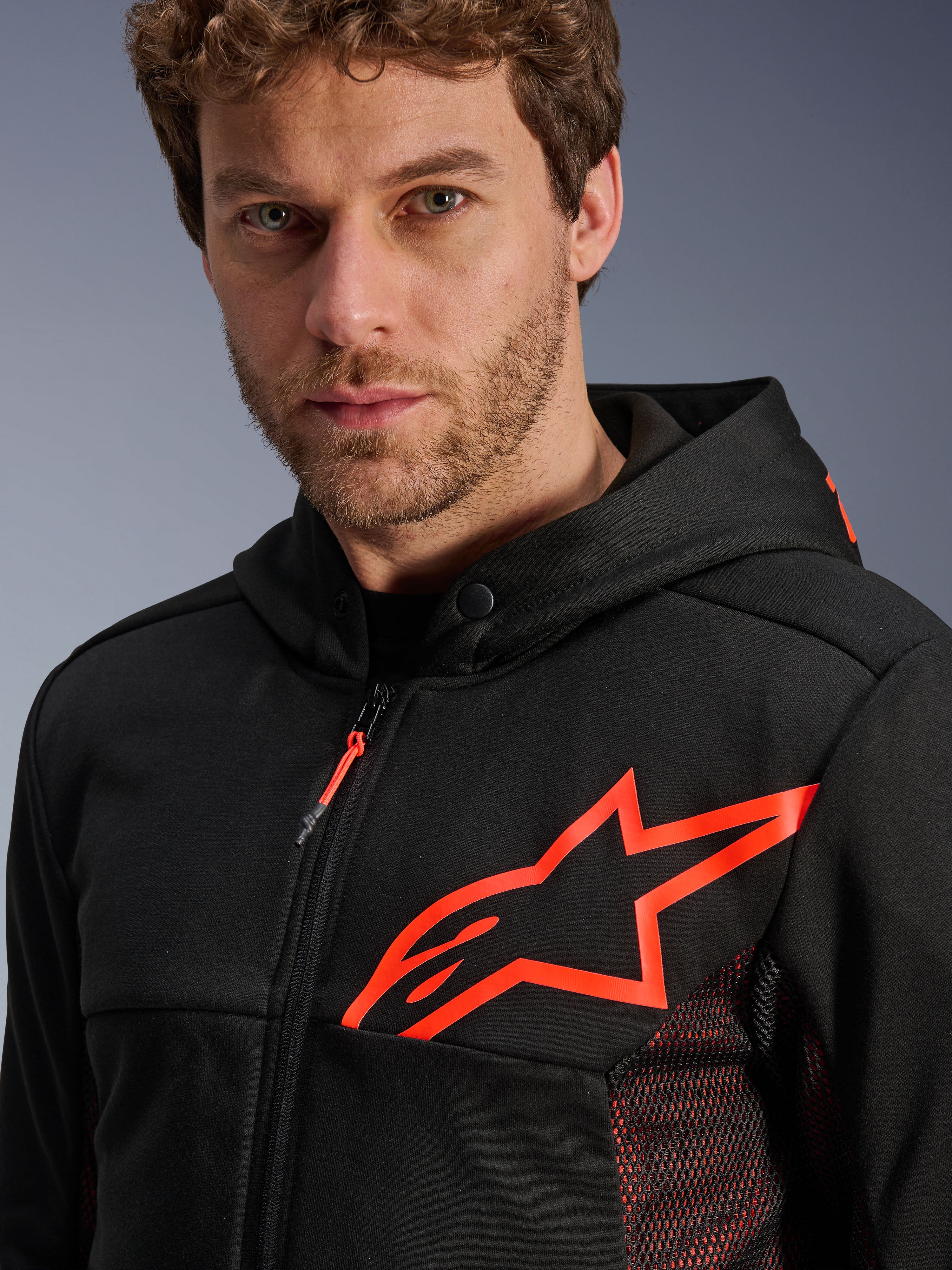 Alpinestars Chrome Air Sport Hoodie, preto e vermelho fluo, grande plano a mostrar o grande logótipo da estrela em vermelho fluo no peito, painéis laterais em malha vermelha para ventilação, fecho de correr frontal e gola com botão de pressão, equipamento de motociclismo urbano casual