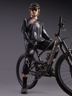 Jersey Alpinestars A-Dura Ride de mangas compridas, camuflado preto e logótipo laranja, usada por ciclista em estúdio.
