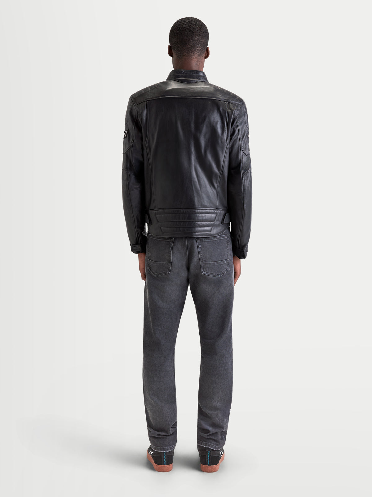 Alpinestars Revern casaco de couro, casaco de moto urbano contemporâneo para homem, preto, vista traseira num modelo a usar jeans e sapatos de moto casuais, destacando a construção em couro bovino de flor integral e o estilo biker de herança