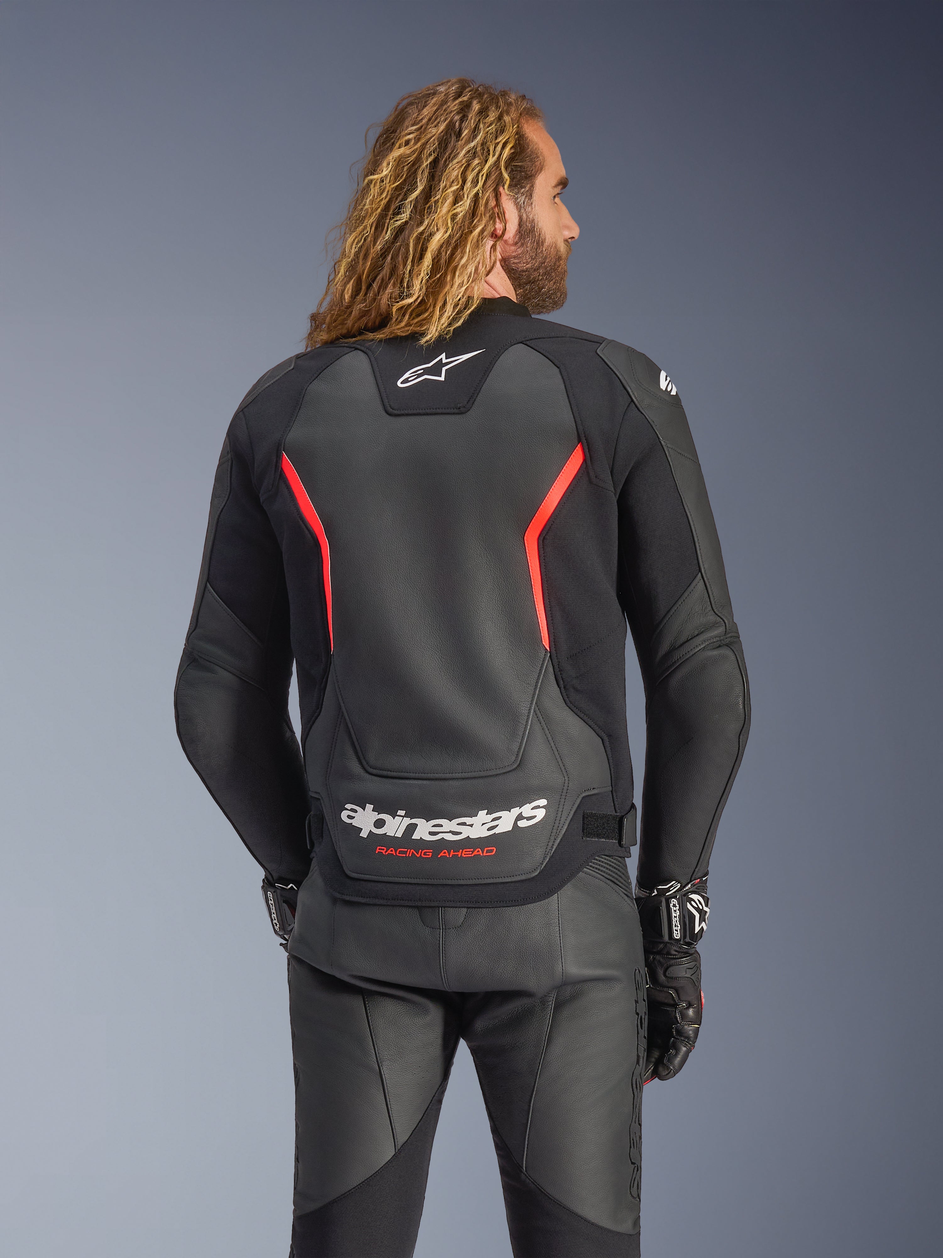 Casaco de Pele Alpinestars GP Force V2, Preto e Vermelho Fluo, vista traseira num modelo, com o logótipo Alpinestars branco e o texto 'RACING AHEAD', combinado com calças e luvas de motociclismo em pele preta.