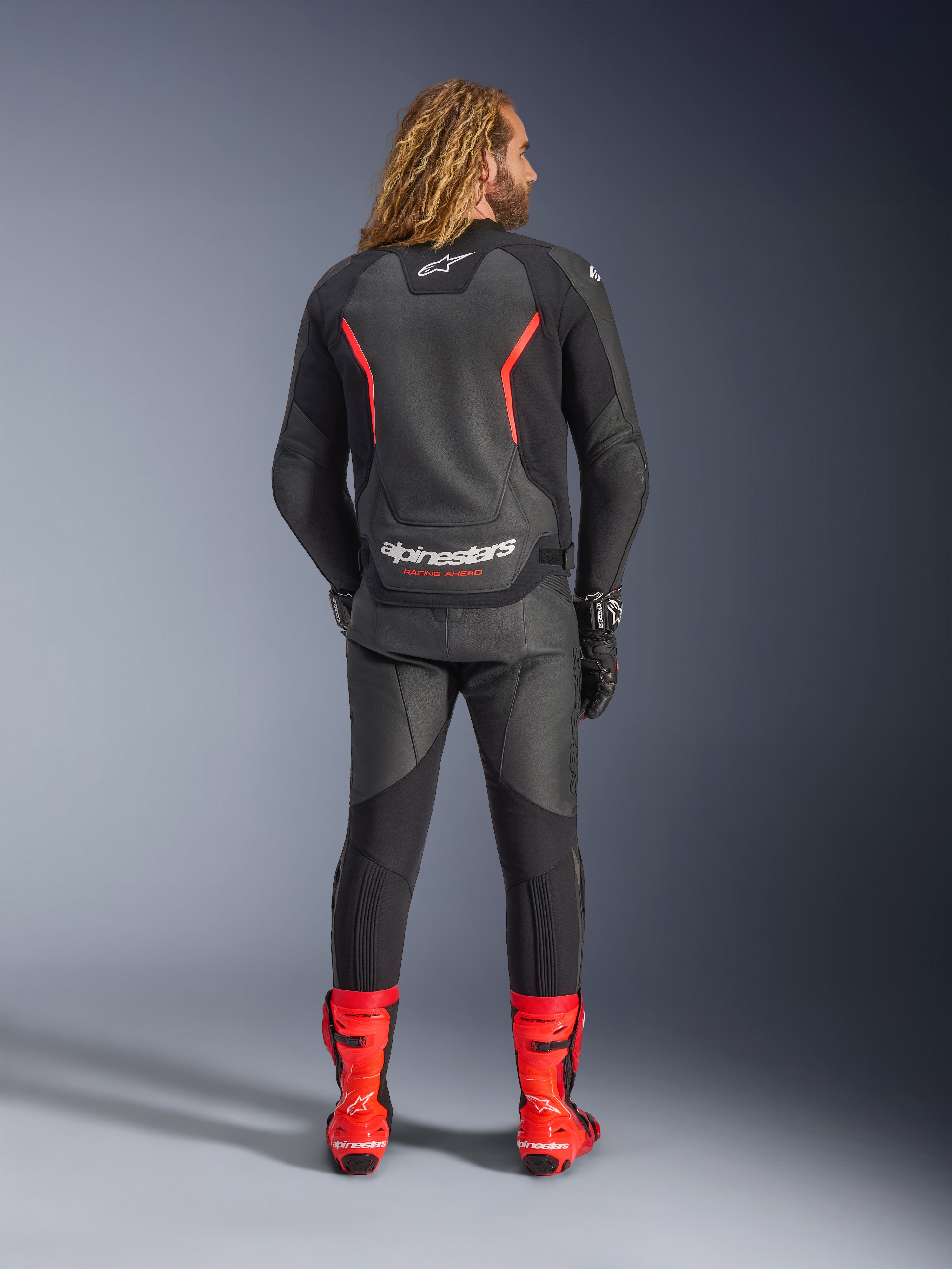 Casaco de Pele Alpinestars GP Force V2, preto e vermelho fluo, visto de costas, usado com calças de pele pretas a condizer e botas de corrida vermelhas vibrantes, apresentando linhas de destaque vermelhas e logótipo Alpinestars em branco nas costas, equipamento profissional de motociclismo desportivo.