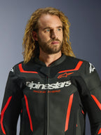 Casaco de Pele Alpinestars GP Force V2 em preto e vermelho fluo, usado por um modelo masculino com cabelo loiro comprido e encaracolado, com a marca Alpinestars em destaque a branco e o texto 'RACING AHEAD' no peito; casaco de motociclismo de pele premium concebido para performance desportiva.