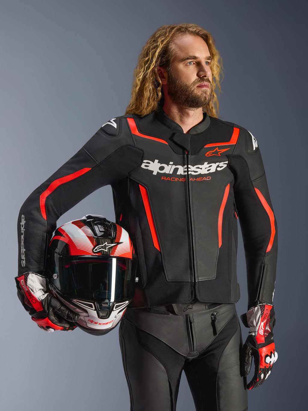 Casaco de pele Alpinestars GP Force V2, preto e vermelho fluo, usado por um modelo masculino a segurar um capacete a condizer, com logótipos Alpinestars brancos e detalhes em vermelho, combinado com calças de pele e luvas de corrida, equipamento de motociclismo desportivo para estrada e pista.