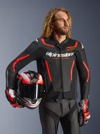 Casaco de pele Alpinestars GP Force V2, preto e vermelho fluo, usado por um modelo masculino a segurar um capacete a condizer, com logótipos Alpinestars brancos e detalhes em vermelho, combinado com calças de pele e luvas de corrida, equipamento de motociclismo desportivo para estrada e pista.