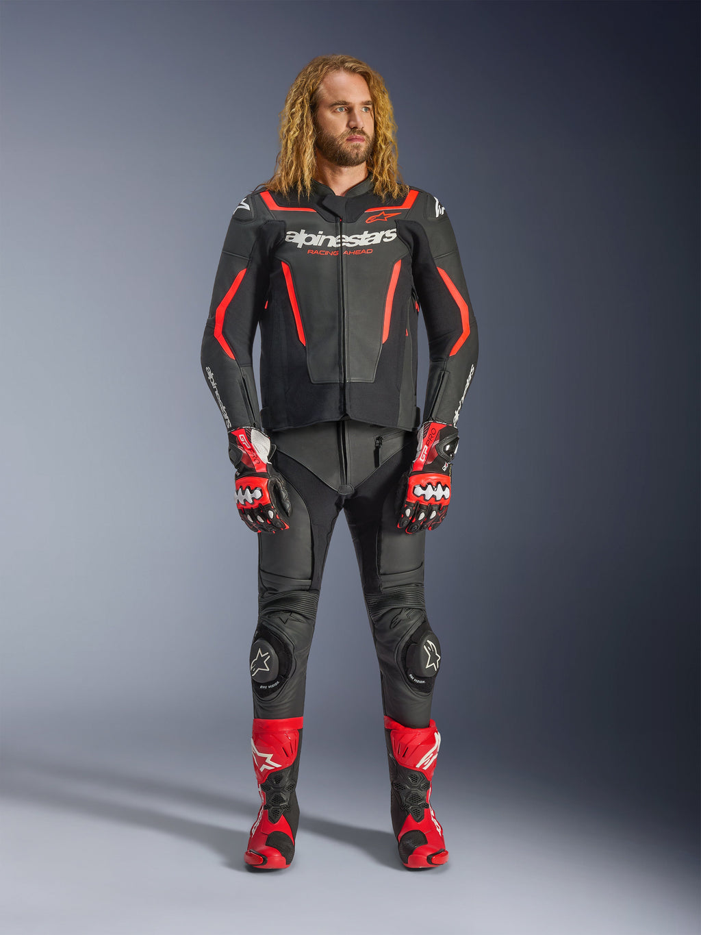 Modelo masculino a usar o casaco de pele Alpinestars GP Force V2 em preto e vermelho flúor, combinado com calças de pele a condizer, luvas de corrida vermelhas e botas de performance vermelhas, equipamento desportivo profissional para motociclismo