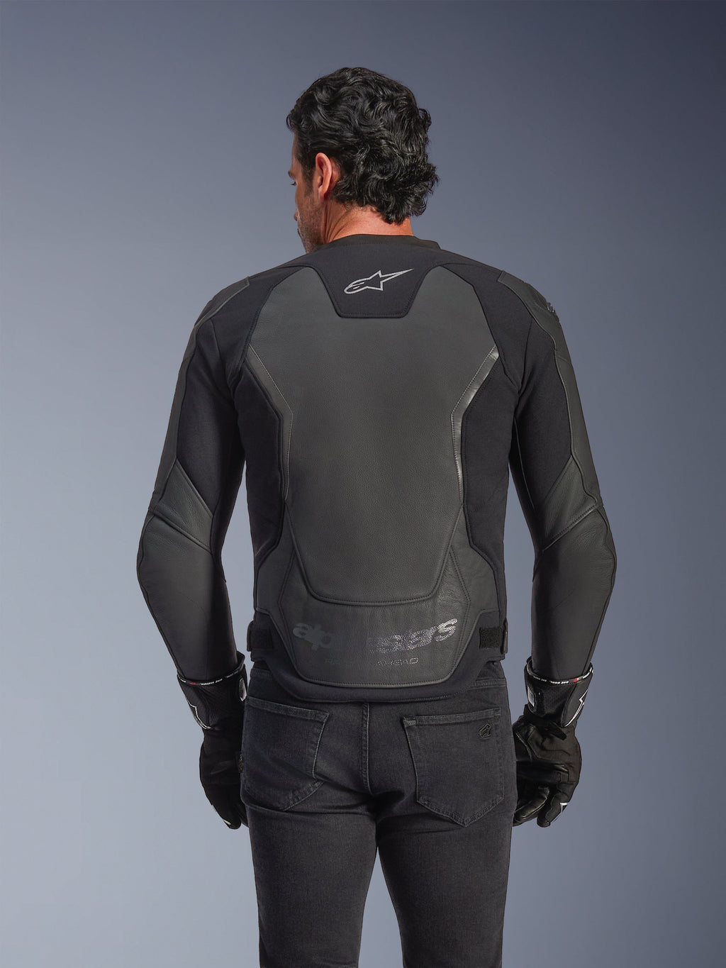 Casaco de Pele Alpinestars GP Force V2, Preto, vista traseira de um homem a usar o casaco com luvas pretas e calças de ganga, com grandes painéis de proteção em pele nas costas, logótipo Alpinestars na parte superior das costas e marca gravada em relevo na cintura, equipamento de motociclismo de inspiração desportiva