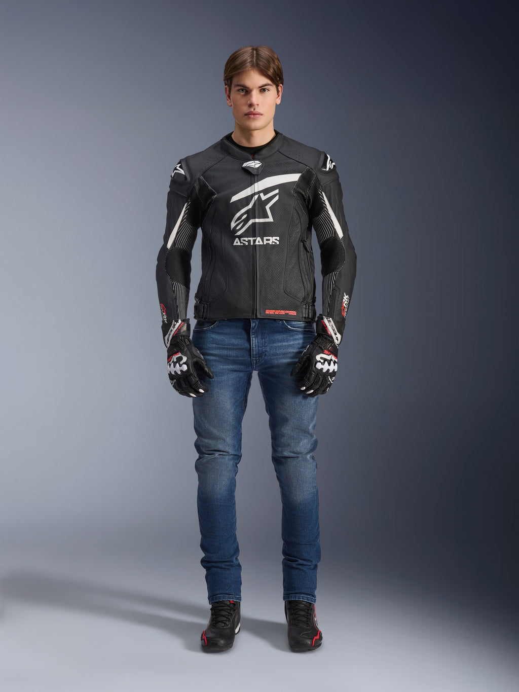 Modelo masculino a usar a Jaqueta de Couro Alpinestars GP Plus R V4 Rideknit em preto e branco, acompanhado por calças de ganga de motociclismo azuis, luvas de motociclismo pretas e sapatos de motociclismo pretos, apresentando um grande logótipo Astars branco no peito, painéis de couro perfurado e riscas de corrida brancas nas mangas, vista de corpo inteiro em pé