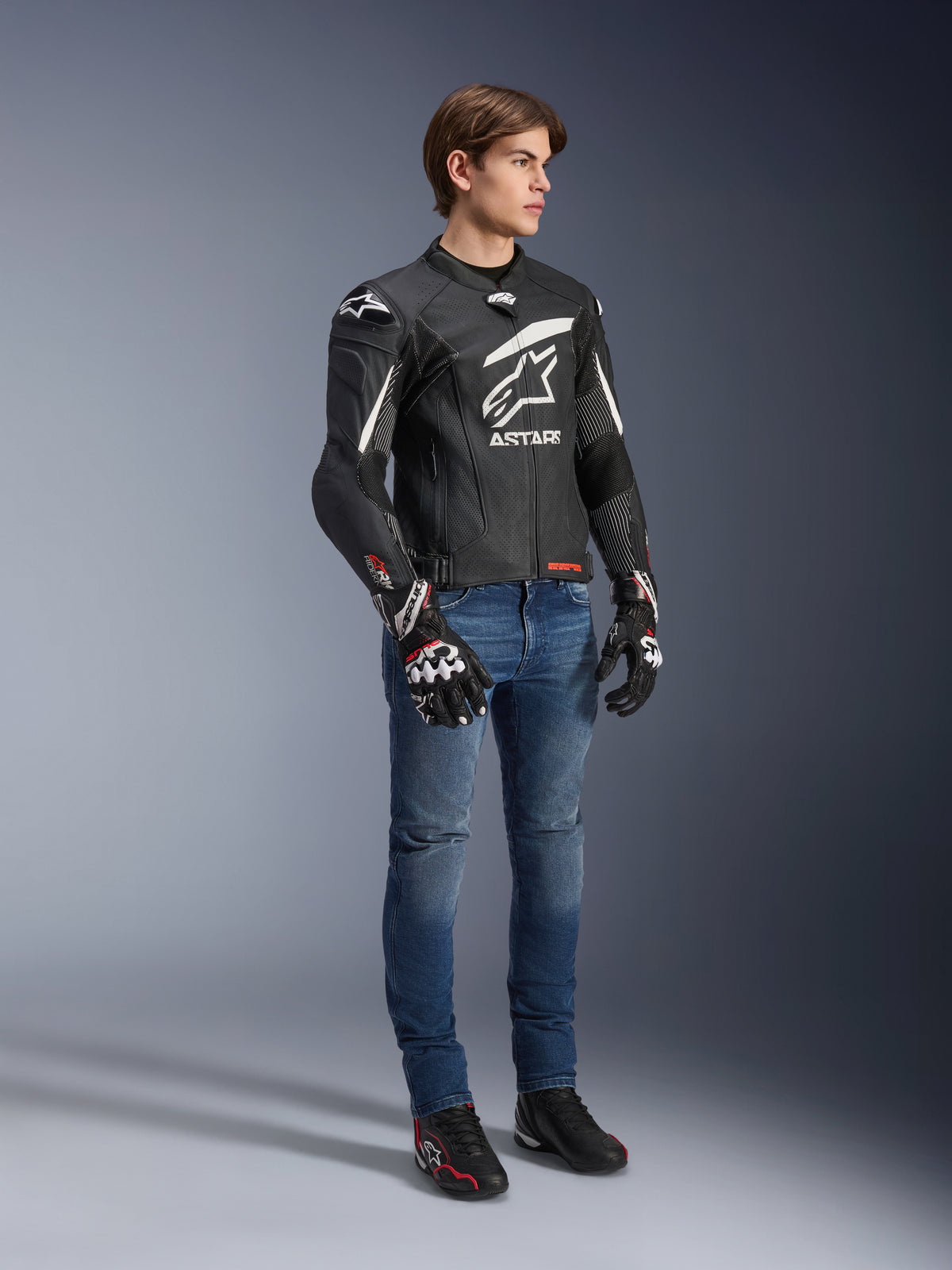 Alpinestars GP Plus R V4 Rideknit Leather Jacket, preto e branco, casaco de motociclismo em couro usado por um modelo com calças de ganga azuis e luvas pretas, com um logótipo Astars branco proeminente no peito, painéis perfurados e detalhes brancos nos ombros.