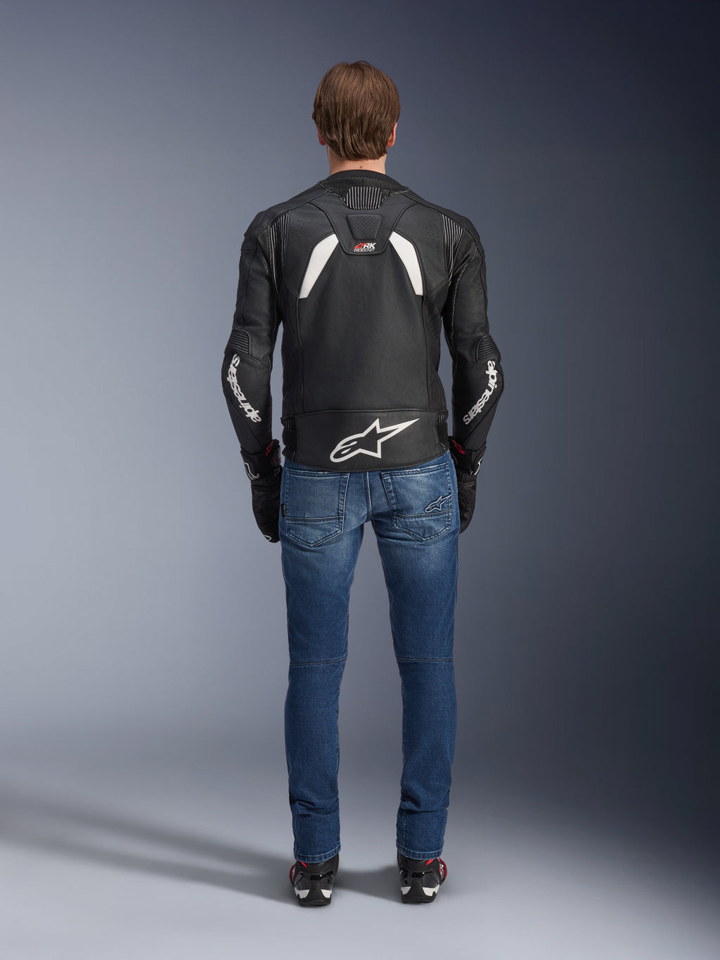 Vista traseira do Casaco de Couro Alpinestars GP Plus R V4 Rideknit, preto e branco, com um logótipo branco grande na parte inferior das costas e marca nas mangas, usado com calças de ganga de motociclismo azuis e botas pretas, realçando a construção técnica e o ajuste desportivo.