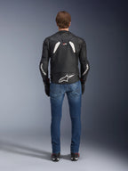 Vista traseira do Casaco de Couro Alpinestars GP Plus R V4 Rideknit, preto e branco, com um logótipo branco grande na parte inferior das costas e marca nas mangas, usado com calças de ganga de motociclismo azuis e botas pretas, realçando a construção técnica e o ajuste desportivo.