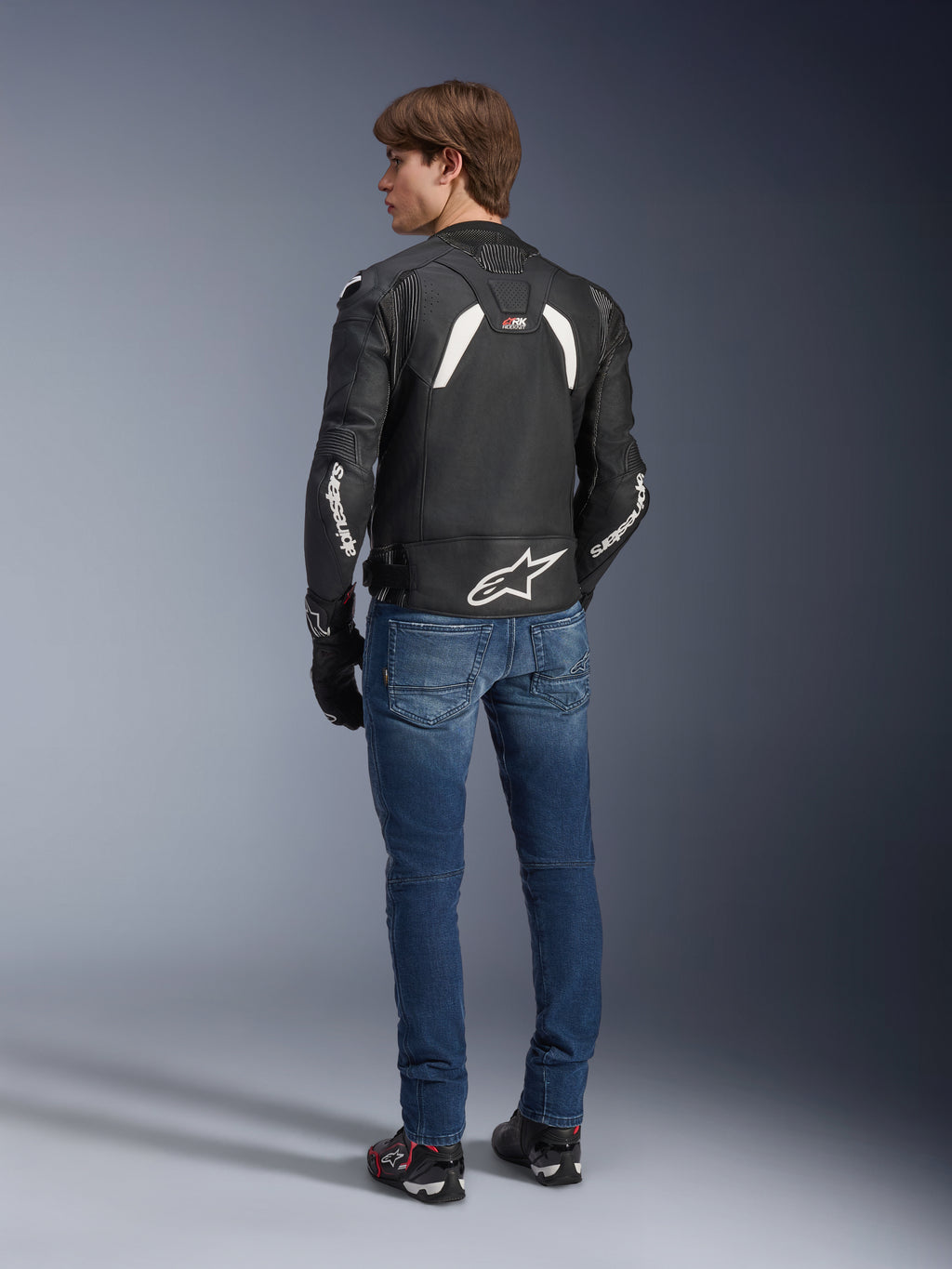 Casaco de couro Alpinestars GP Plus R V4 Rideknit em preto e branco, vista traseira num modelo, com a marca Alpinestars a branco nas mangas e um logótipo de estrela grande na parte inferior das costas, usado com calças de ganga azuis e sapatos de condução pretos, equipamento técnico de motociclismo