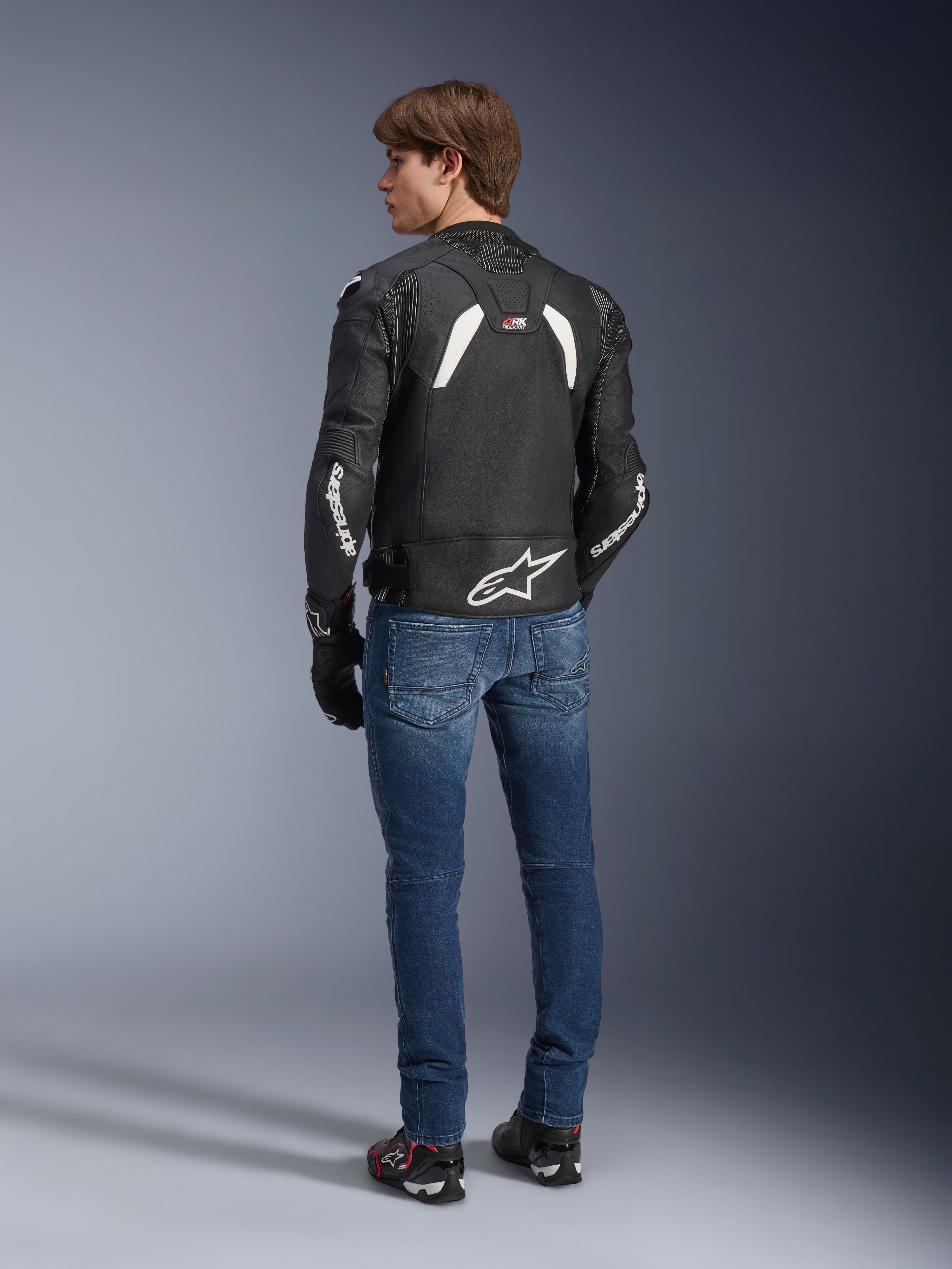 Casaco de couro Alpinestars GP Plus R V4 Rideknit em preto e branco, vista traseira num modelo, com a marca Alpinestars a branco nas mangas e um logótipo de estrela grande na parte inferior das costas, usado com calças de ganga azuis e sapatos de condução pretos, equipamento técnico de motociclismo