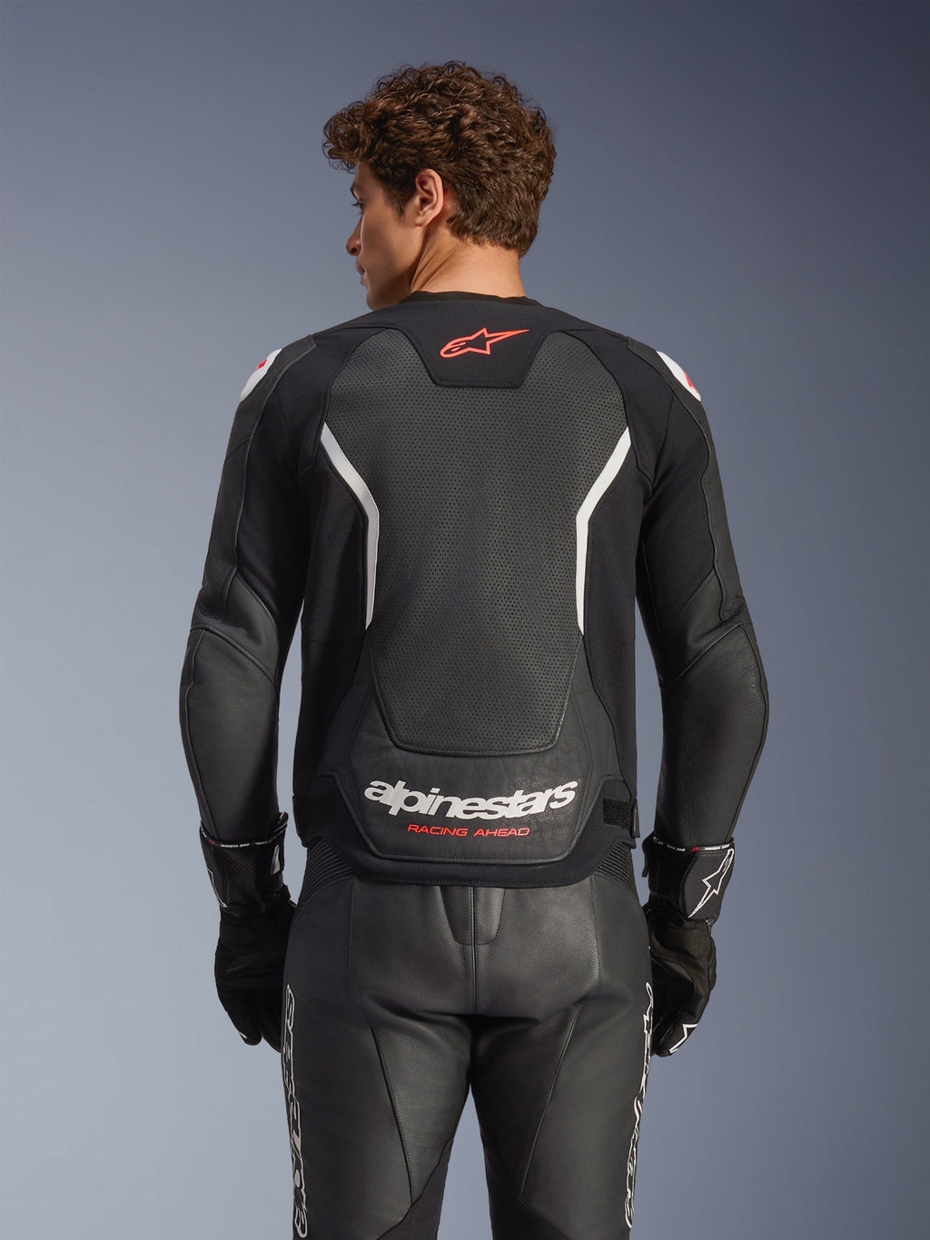 Alpinestars GP Force V2 Air Leather Jacket, Black with White and Red accents, rear view showing perforated leather back panel, white Alpinestars branding, and red logo details, worn with matching black leather racing pants and gloves.