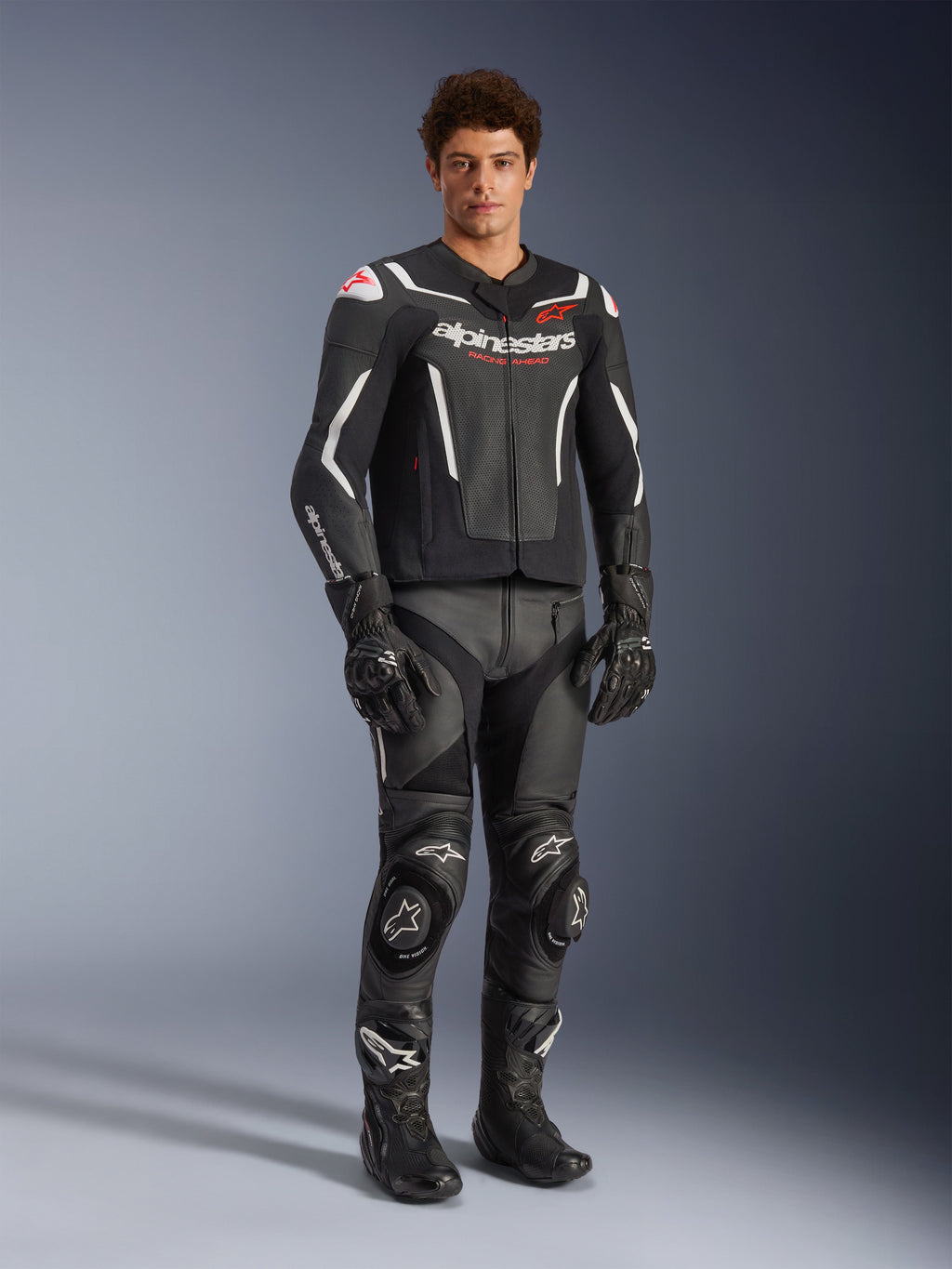 Alpinestars GP Force V2 Air Leather Jacket in Black and White, worn by a male rider with matching black racing pants, gloves, and boots, featuring perforated leather panels, white accents, and red Alpinestars logo on the chest, sport motorcycle riding gear