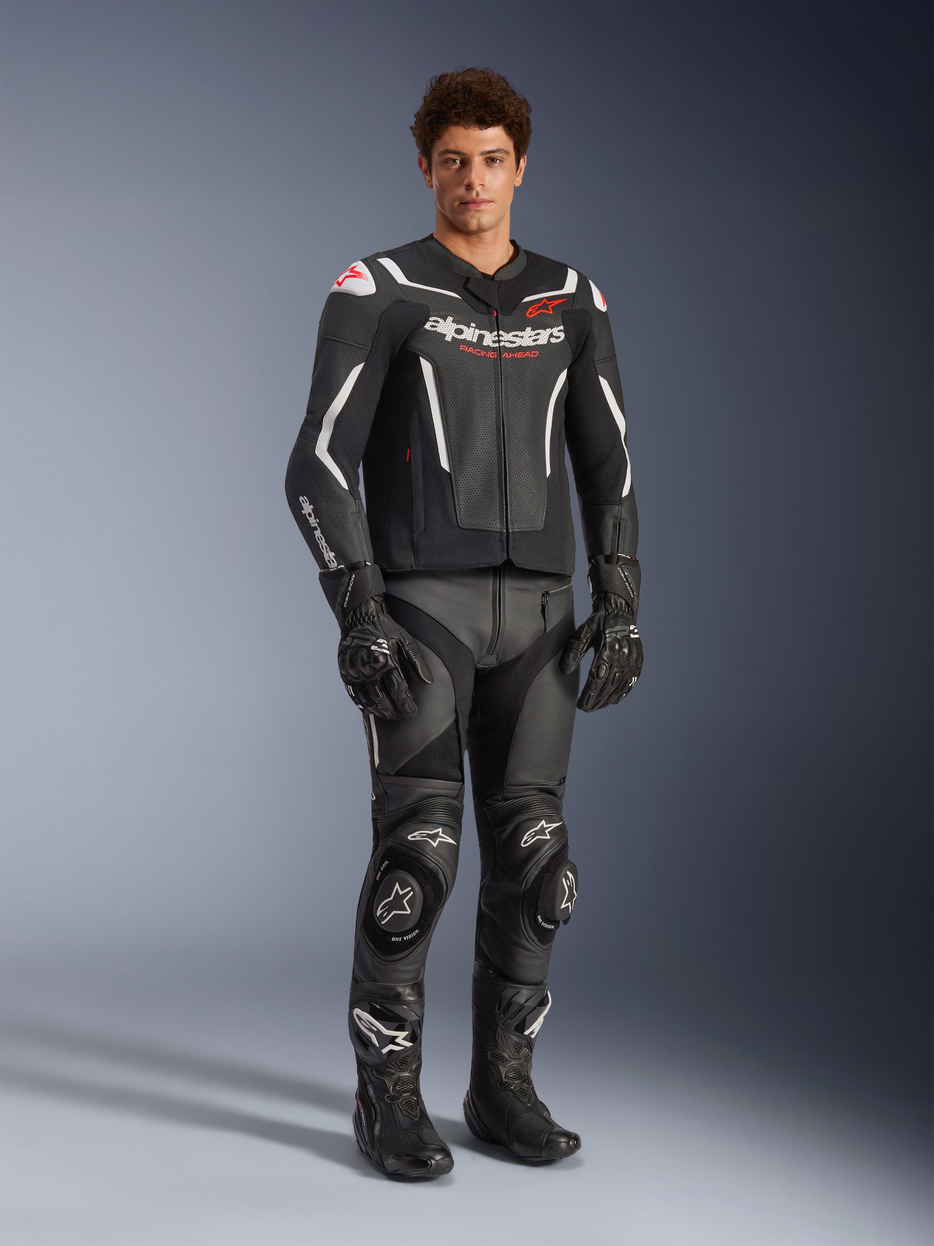 Alpinestars GP Force V2 Air Leather Jacket in Black and White, worn by a male rider with matching black racing pants, gloves, and boots, featuring perforated leather panels, white accents, and red Alpinestars logo on the chest, sport motorcycle riding gear