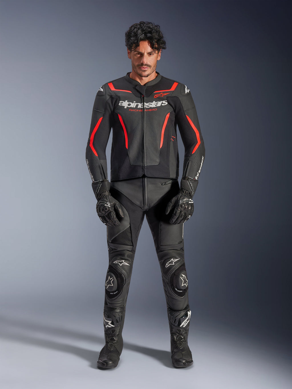 Male model wearing Alpinestars GP Force V2 Air Leather Jacket in Black and Red Fluo, featuring perforated chest panels and white branding, paired with black motorcycle leather pants, black gloves, and black boots for a complete racing look.