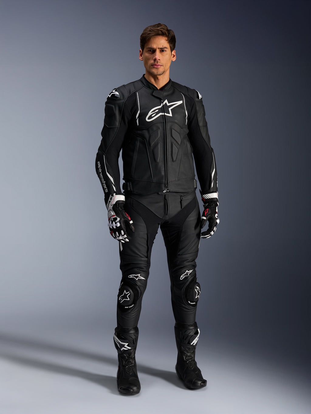Modelo masculino a usar o casaco de pele Alpinestars Dusk em preto e branco, com logótipos brancos proeminentes no peito e nos ombros, combinado com calças de motociclismo em pele pretas a condizer, luvas pretas e brancas e botas pretas, equipamento de corrida desportivo completo para uso em estrada