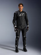 Modelo masculino a usar o casaco de pele Alpinestars Dusk em preto e branco, com logótipos brancos proeminentes no peito e nos ombros, combinado com calças de motociclismo em pele pretas a condizer, luvas pretas e brancas e botas pretas, equipamento de corrida desportivo completo para uso em estrada