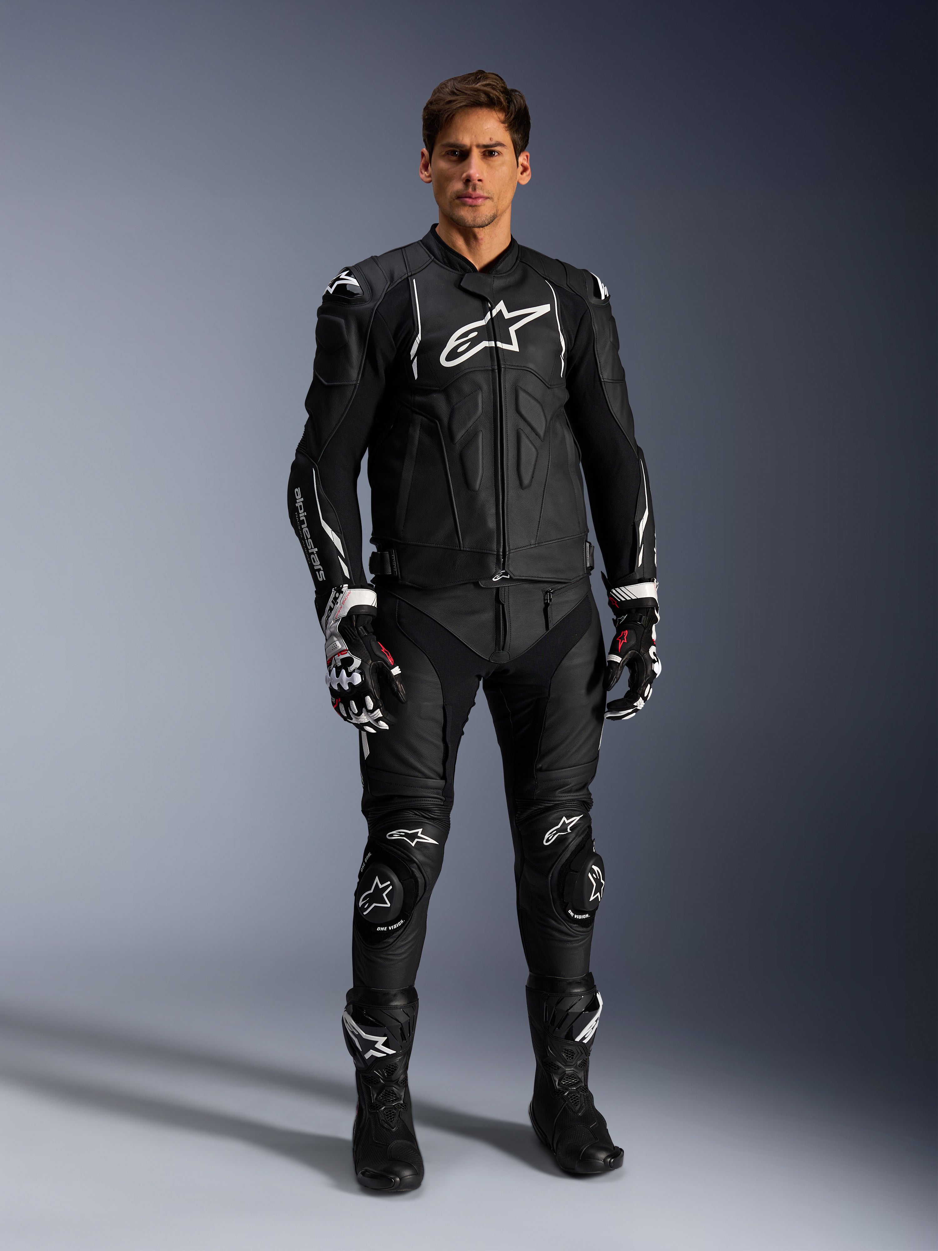 Modelo masculino a usar o casaco de pele Alpinestars Dusk em preto e branco, com logótipos brancos proeminentes no peito e nos ombros, combinado com calças de motociclismo em pele pretas a condizer, luvas pretas e brancas e botas pretas, equipamento de corrida desportivo completo para uso em estrada