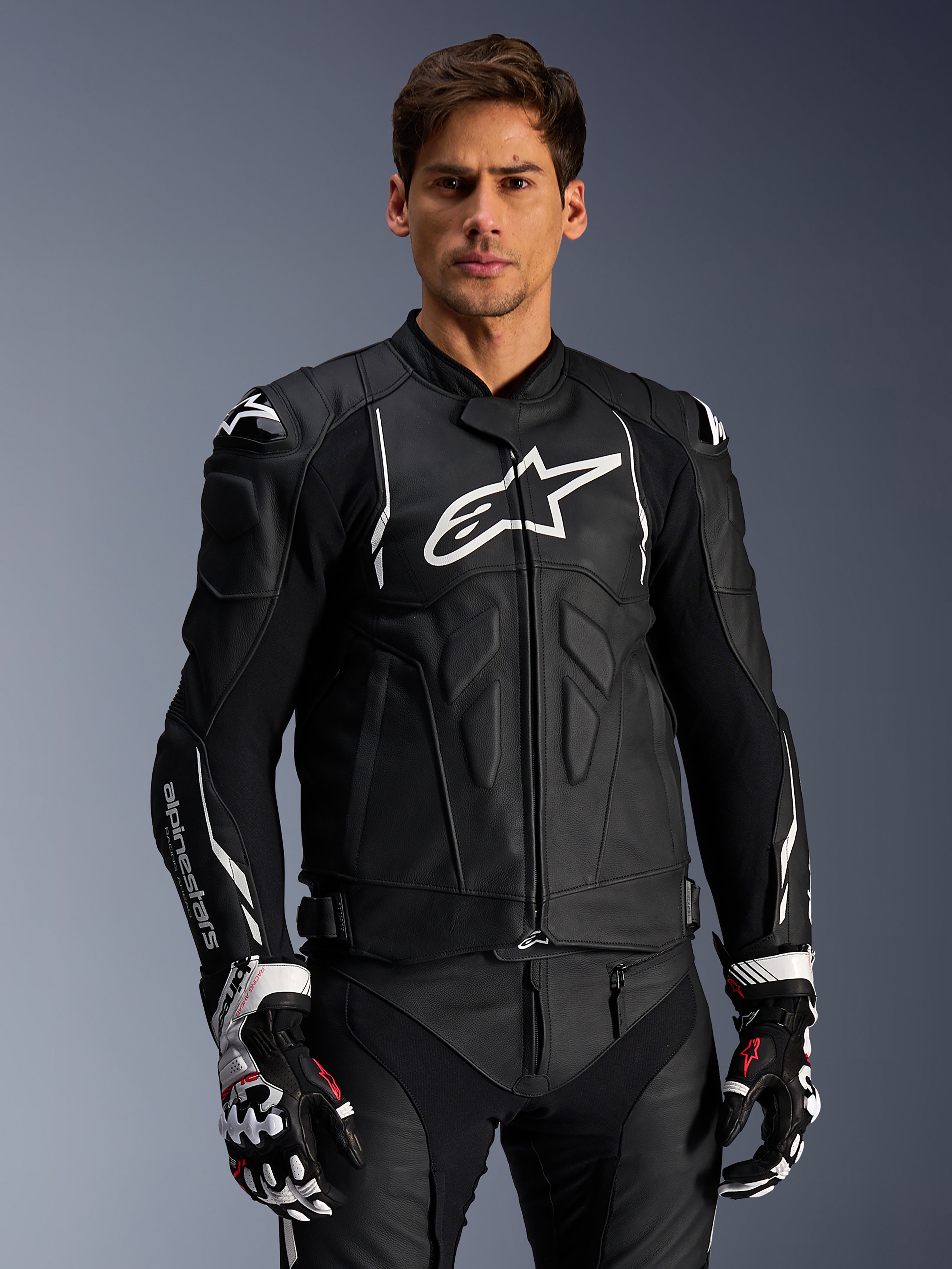Alpinestars Dusk Leather Jacket, preto e branco, casaco de motociclismo em pele com áreas de proteção acolchoadas e logótipos brancos, usado com luvas de corrida pretas e brancas com detalhes em vermelho, equipamento de condução desportiva profissional