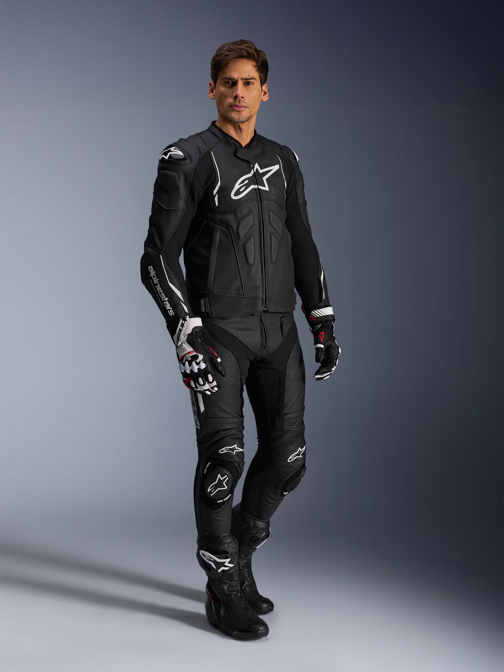 Motociclista com o Casaco de Pele Alpinestars Dusk em preto e branco, casaco de pele premium com logótipo branco de grandes dimensões no peito, combinado com calças de corrida em pele, luvas e botas a condizer, equipamento completo de alto desempenho para condução desportiva