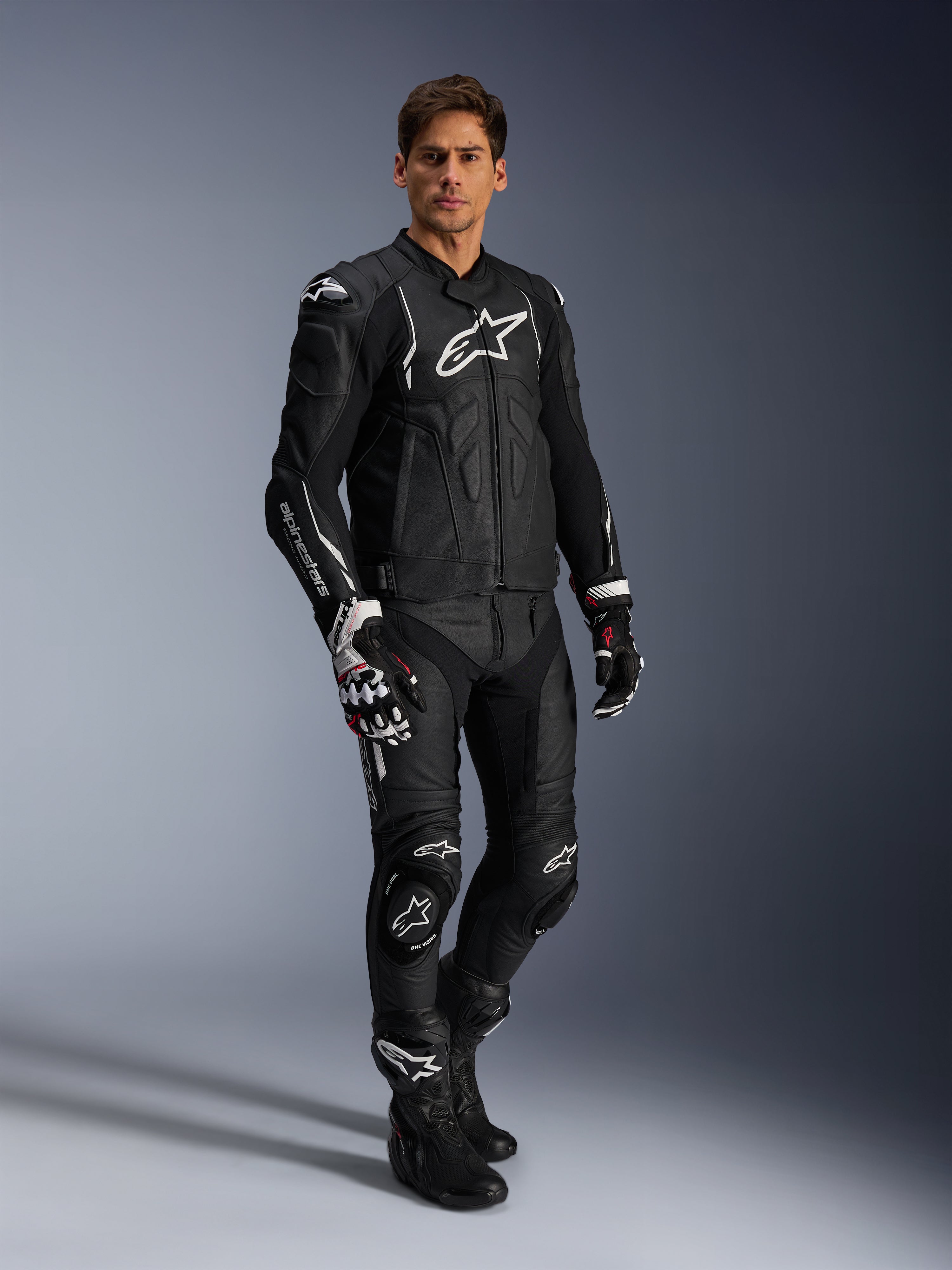 Motociclista com o Casaco de Pele Alpinestars Dusk em preto e branco, casaco de pele premium com logótipo branco de grandes dimensões no peito, combinado com calças de corrida em pele, luvas e botas a condizer, equipamento completo de alto desempenho para condução desportiva