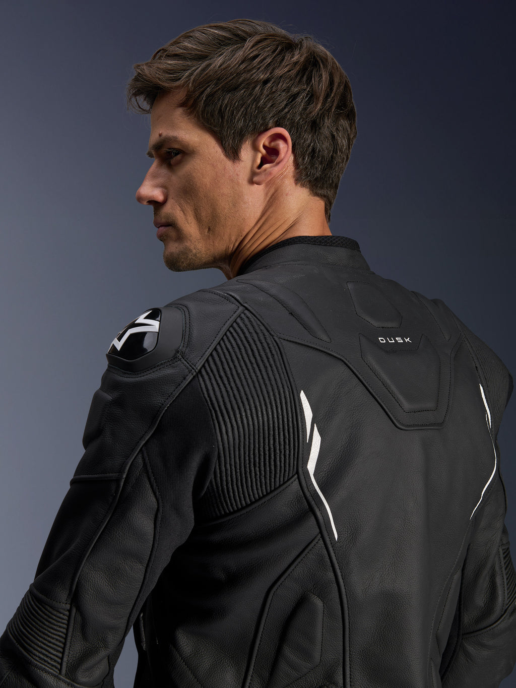Casaco de Pele Alpinestars Dusk, preto com detalhes brancos, vista traseira no modelo, com protetores de ombro DFS com logótipo Alpinestars, painéis elásticos de pele em sanfona para mobilidade e marca 'DUSK' na parte superior das costas, casaco de motociclismo em pele bovina premium para condução em estrada e desportiva