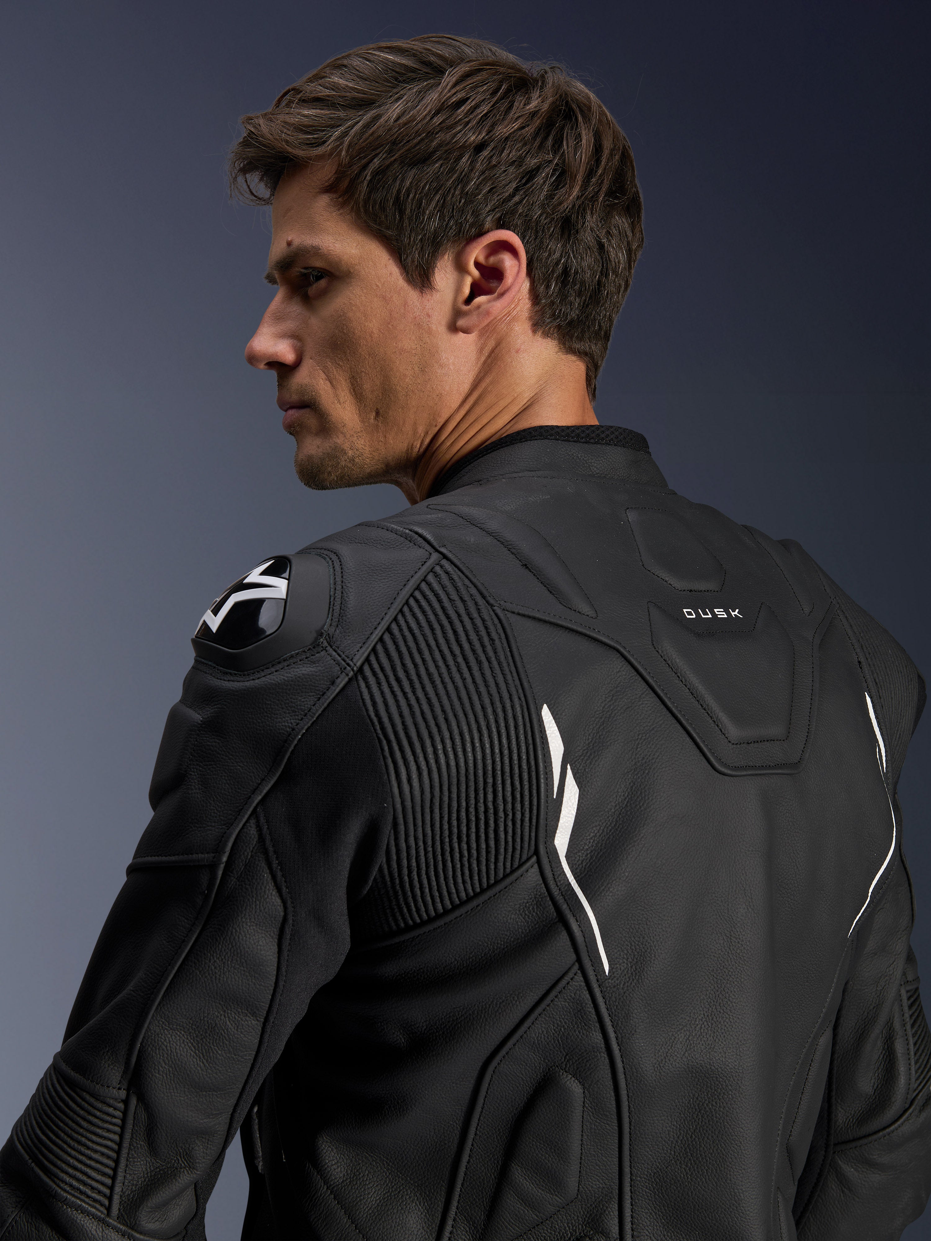 Casaco de Pele Alpinestars Dusk, preto com detalhes brancos, vista traseira no modelo, com protetores de ombro DFS com logótipo Alpinestars, painéis elásticos de pele em sanfona para mobilidade e marca 'DUSK' na parte superior das costas, casaco de motociclismo em pele bovina premium para condução em estrada e desportiva