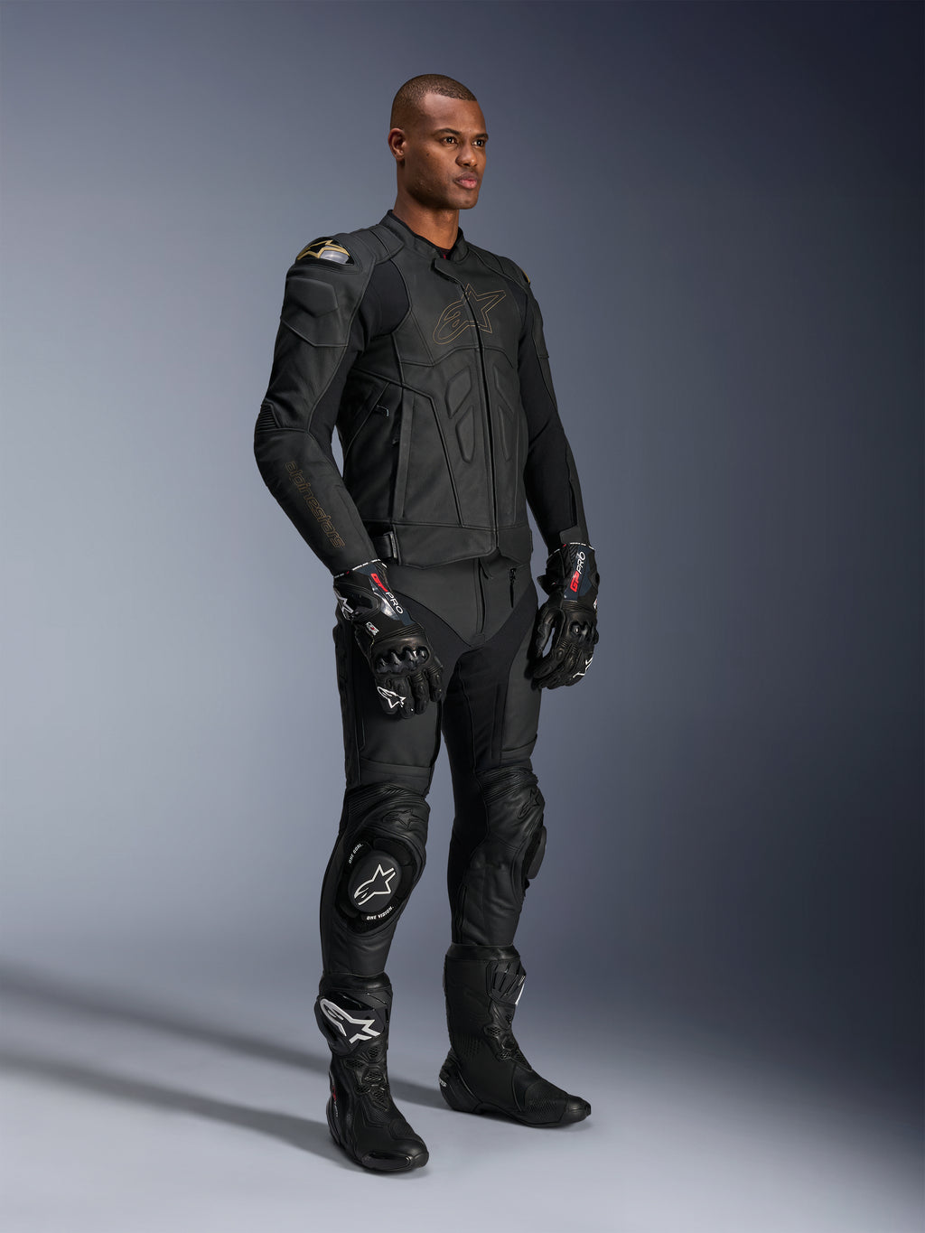 Male model wearing Alpinestars Missile V3 Leather Pants and matching black leather jacket with gold accents, black GP Pro racing gloves, and black racing boots, complete motorcycle racing gear for road use, studio shot.