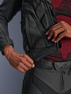 Casaco em Pele Alpinestars Dusk, preto, grande plano do fecho de correr de ligação na cintura a ser fixado a calças de motociclismo em pele, usado com uma camada de base com padrão vermelho e preto, construção em pele resistente para condução em estrada