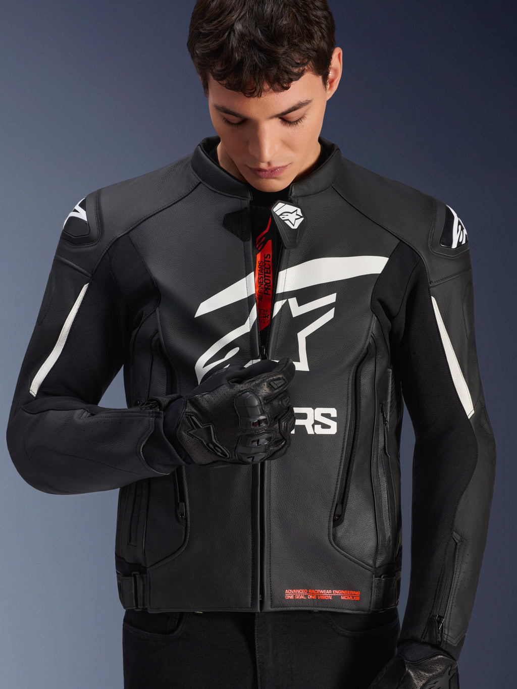 Casaco de Couro Alpinestars GP Plus V4, preto e branco, modelo masculino a fechar o fecho frontal para revelar a marca interior em vermelho, logótipos Alpinestars brancos no peito e ombros, luvas de motociclismo em couro preto, vestuário de condução em couro de inspiração desportiva