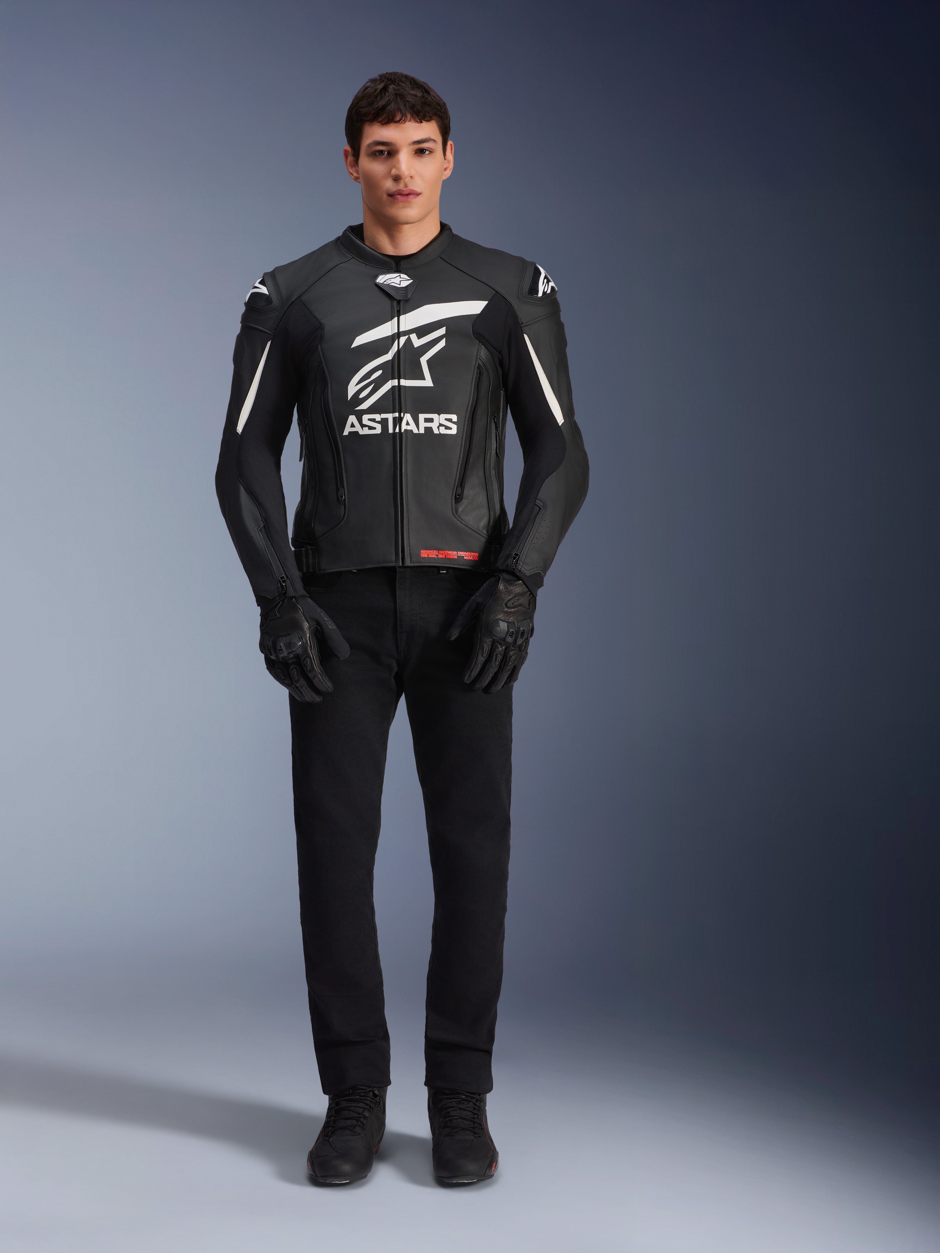 Modelo masculino a usar o Casaco de Couro Alpinestars GP Plus V4, preto e branco, com logótipo Astars em branco no peito e detalhes brancos nos ombros, combinado com luvas e calças pretas, vestuário de motociclismo de estilo desportivo