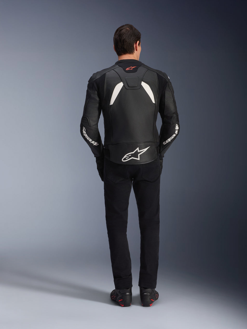 Modelo visto de costas a usar o casaco de couro Alpinestars GP Plus V4 em preto e branco, combinado com calças pretas e botas de motociclismo, com painéis brancos nos ombros e o logótipo Alpinestars nas mangas e na parte inferior das costas.