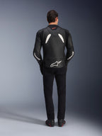 Modelo visto de costas a usar o casaco de couro Alpinestars GP Plus V4 em preto e branco, combinado com calças pretas e botas de motociclismo, com painéis brancos nos ombros e o logótipo Alpinestars nas mangas e na parte inferior das costas.