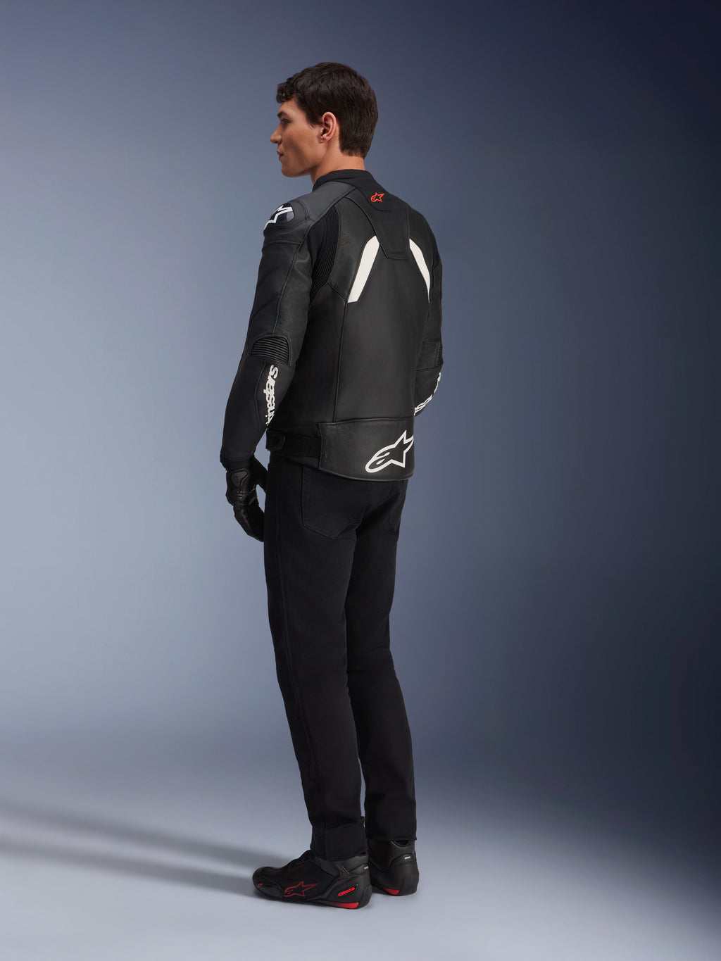 Alpinestars GP Plus V4 Leather Jacket, casaco de couro desportivo, preto e branco, usado por um modelo numa vista de perfil lateral e traseiro, apresentando logótipos Alpinestars brancos na parte inferior das costas e na manga, combinado com calças de motociclismo pretas e botas técnicas para um visual de performance desportiva.