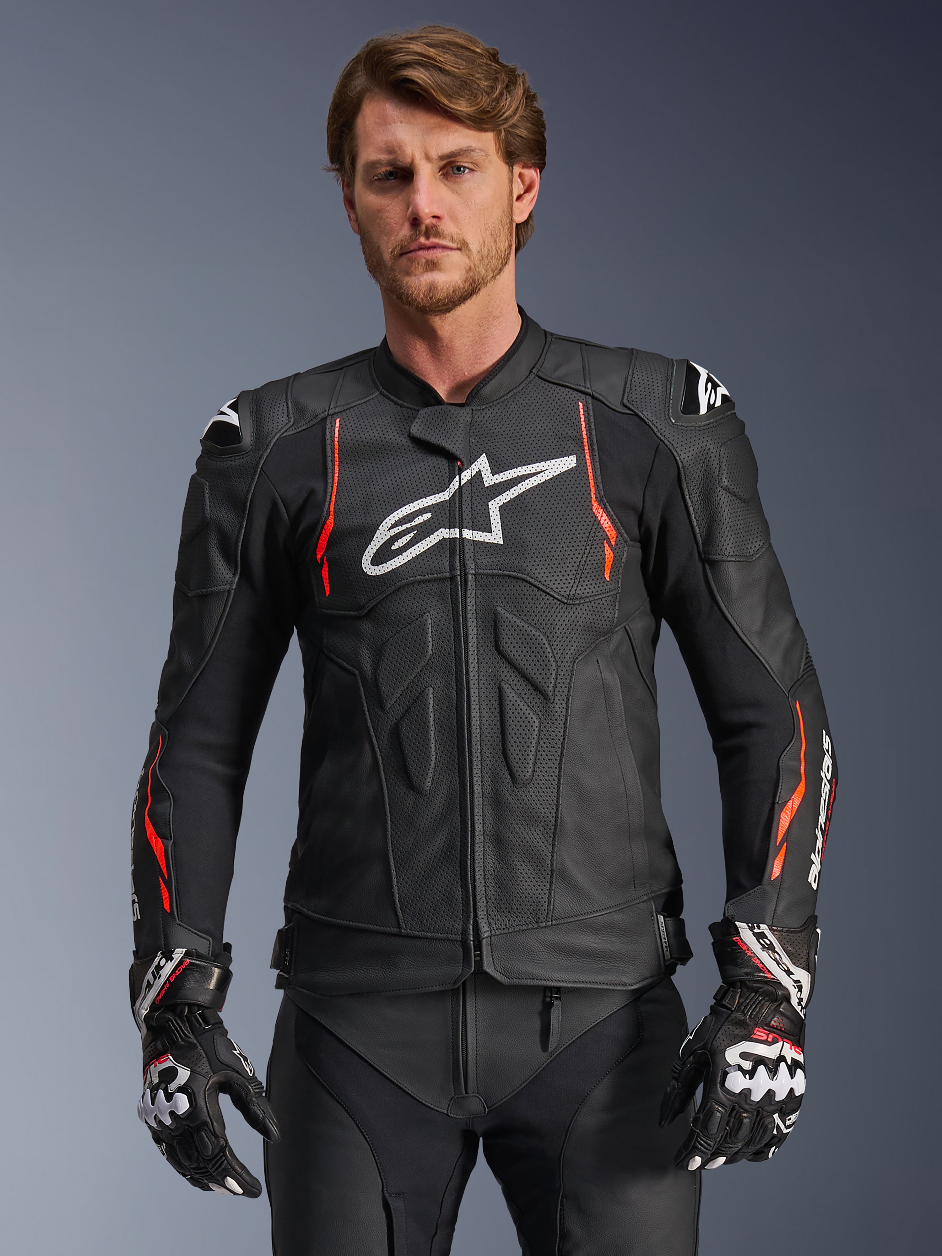 Alpinestars Dusk Airflow Leather Jacket, preto com detalhes em vermelho fluo e branco, casaco de pele para motociclismo de competição, com perfurações extensas para ventilação, logótipo branco no peito e proteções externas nos ombros, usado por um modelo com luvas de corrida a condizer