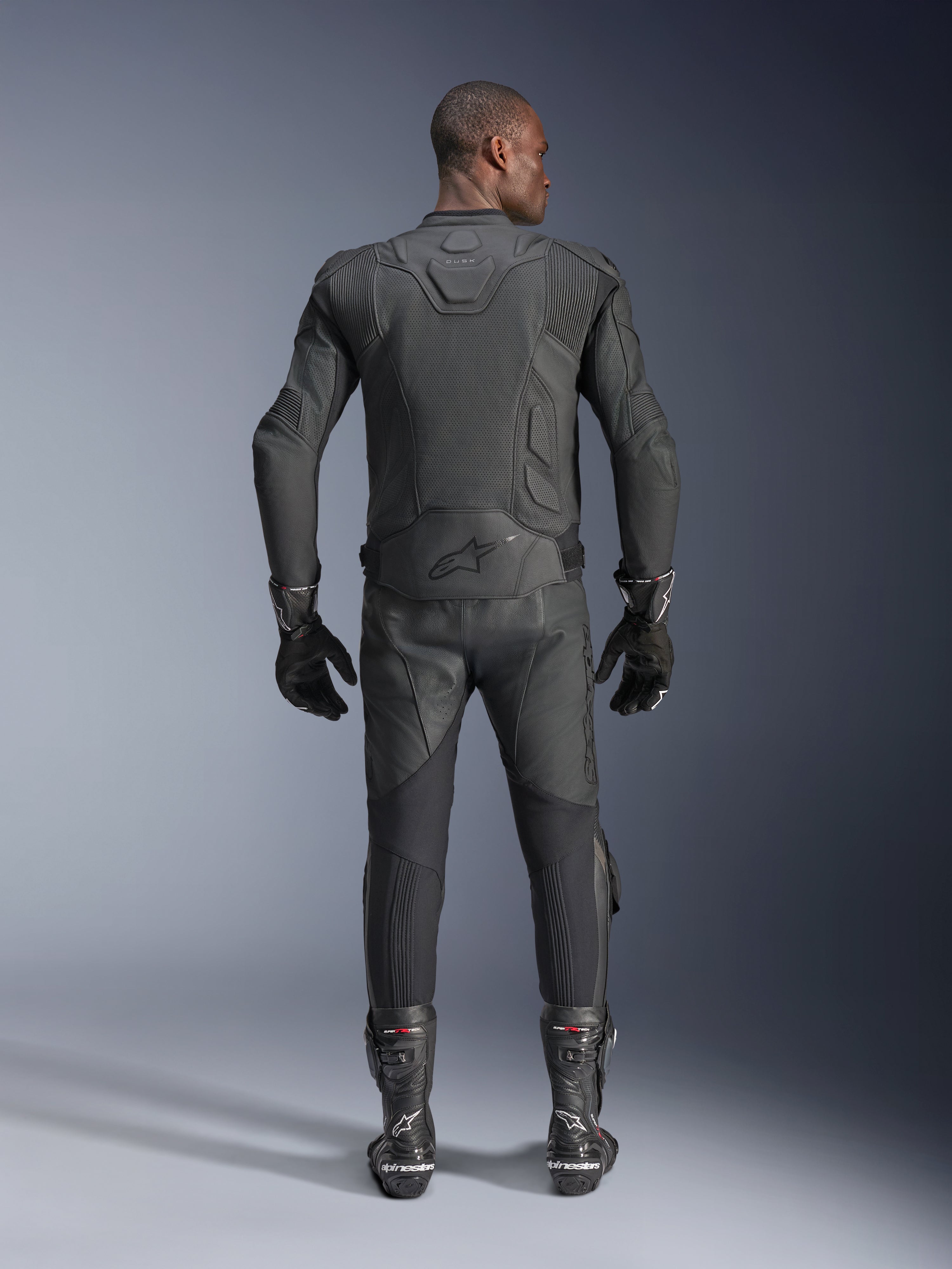Male model shown from the back wearing the Alpinestars Dusk Airflow Leather Jacket in Black, paired with matching black leather racing pants, gloves, and boots, featuring extensive perforations for ventilation and the Alpinestars logo on the lower back.