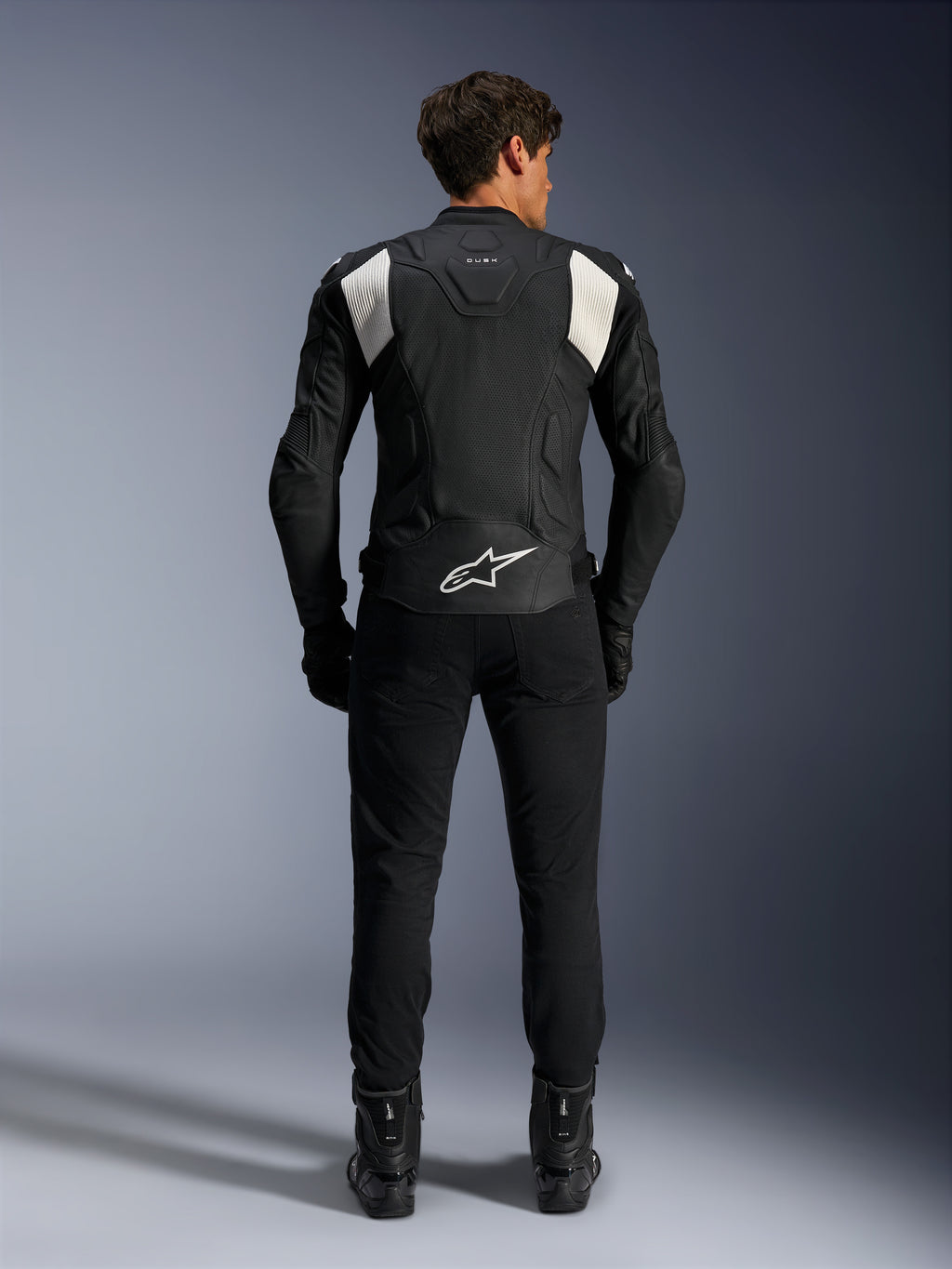 Casaco de Couro Alpinestars Dusk Airflow, Preto e Branco, vista traseira de um piloto a usar calças e botas pretas, com couro perfurado para ventilação, painéis de acordeão brancos nos ombros e logótipo branco na parte inferior das costas