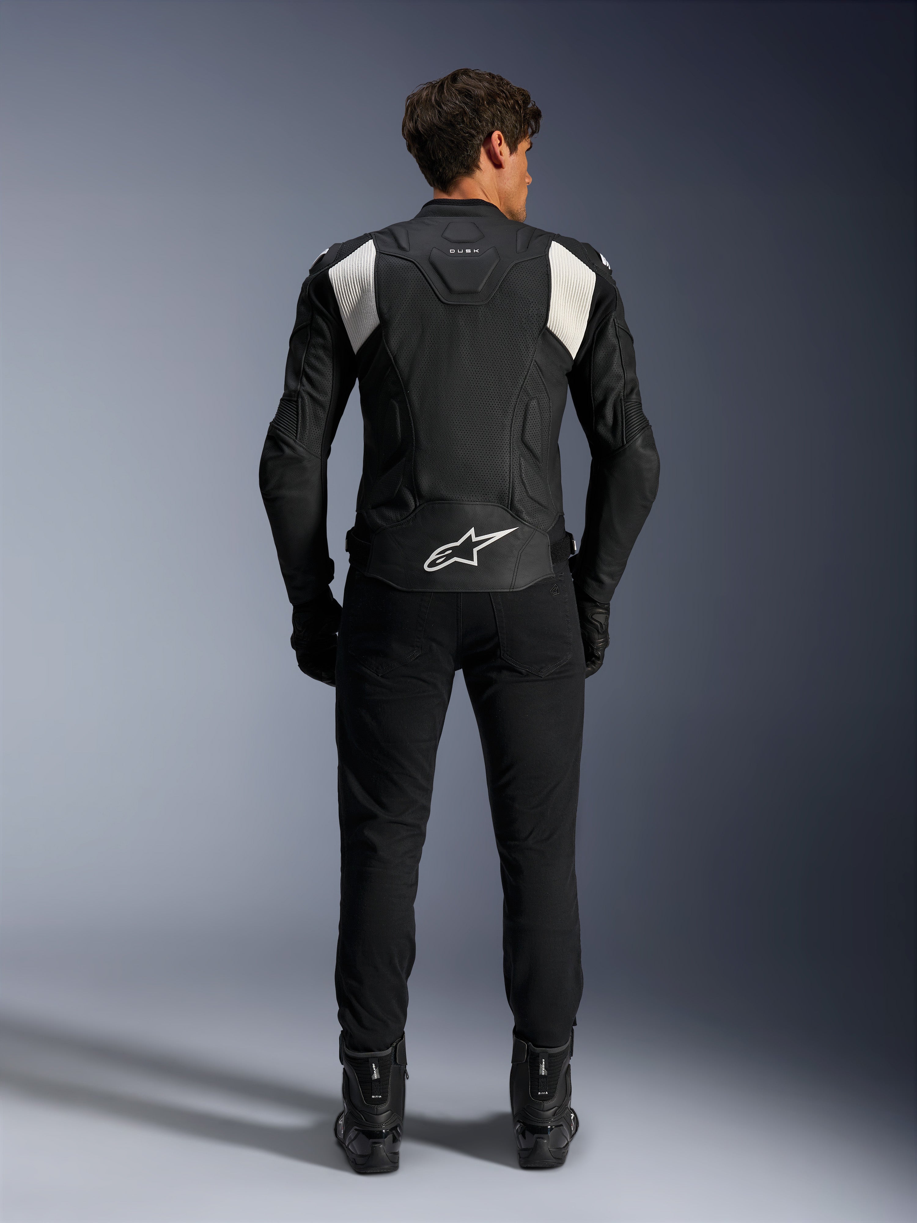 Casaco de Couro Alpinestars Dusk Airflow, Preto e Branco, vista traseira de um piloto a usar calças e botas pretas, com couro perfurado para ventilação, painéis de acordeão brancos nos ombros e logótipo branco na parte inferior das costas