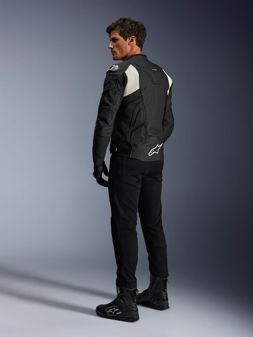 Alpinestars Dusk Airflow Leather Jacket, Preto e Branco, vista traseira de modelo a usar o casaco de couro perfurado com calças e botas pretas a condizer, apresentando painéis em acordeão brancos e logótipo proeminente na parte inferior das costas, equipamento de condução de motociclismo desportivo