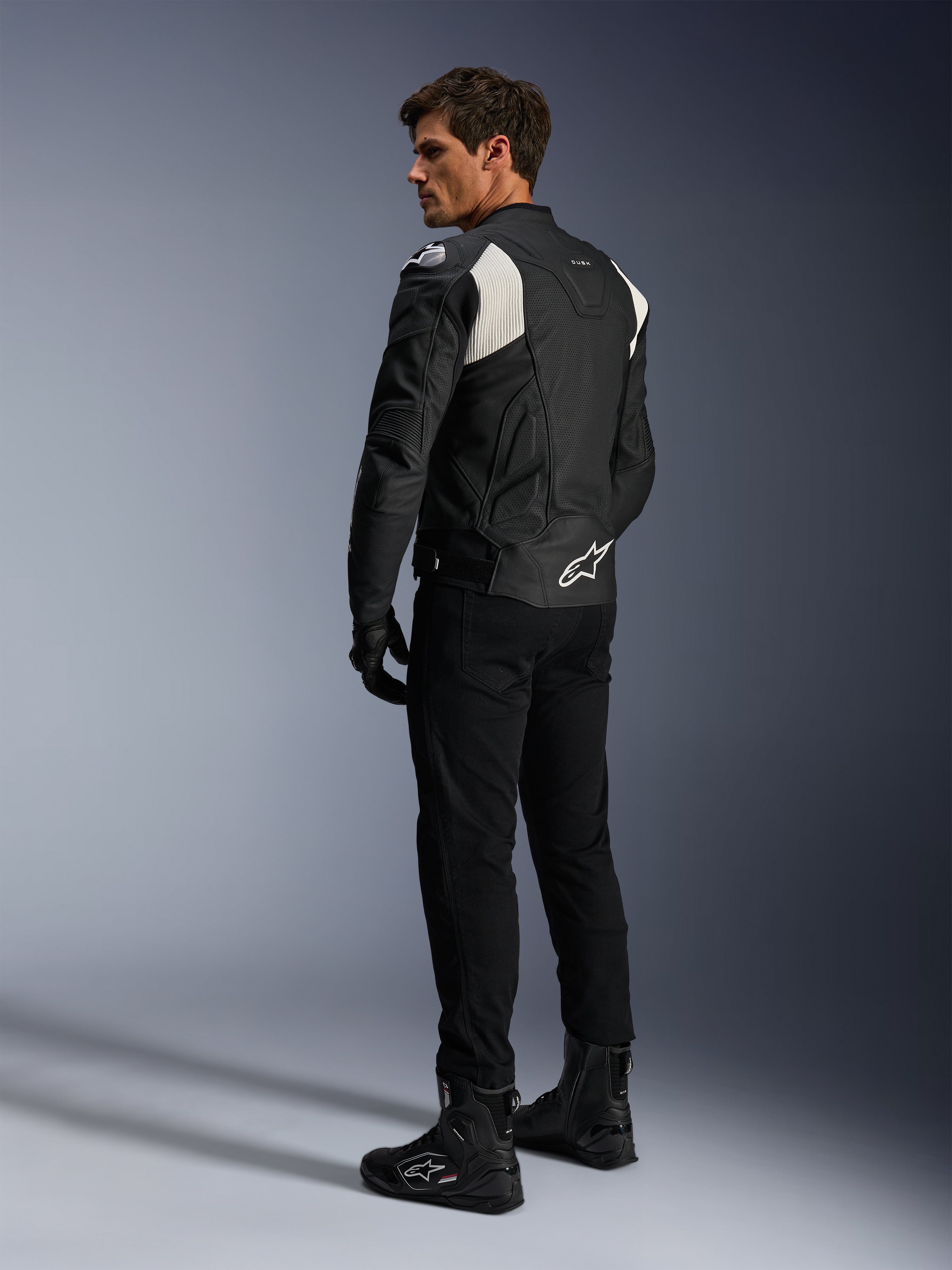 Alpinestars Dusk Airflow Leather Jacket, Preto e Branco, vista traseira de modelo a usar o casaco de couro perfurado com calças e botas pretas a condizer, apresentando painéis em acordeão brancos e logótipo proeminente na parte inferior das costas, equipamento de condução de motociclismo desportivo