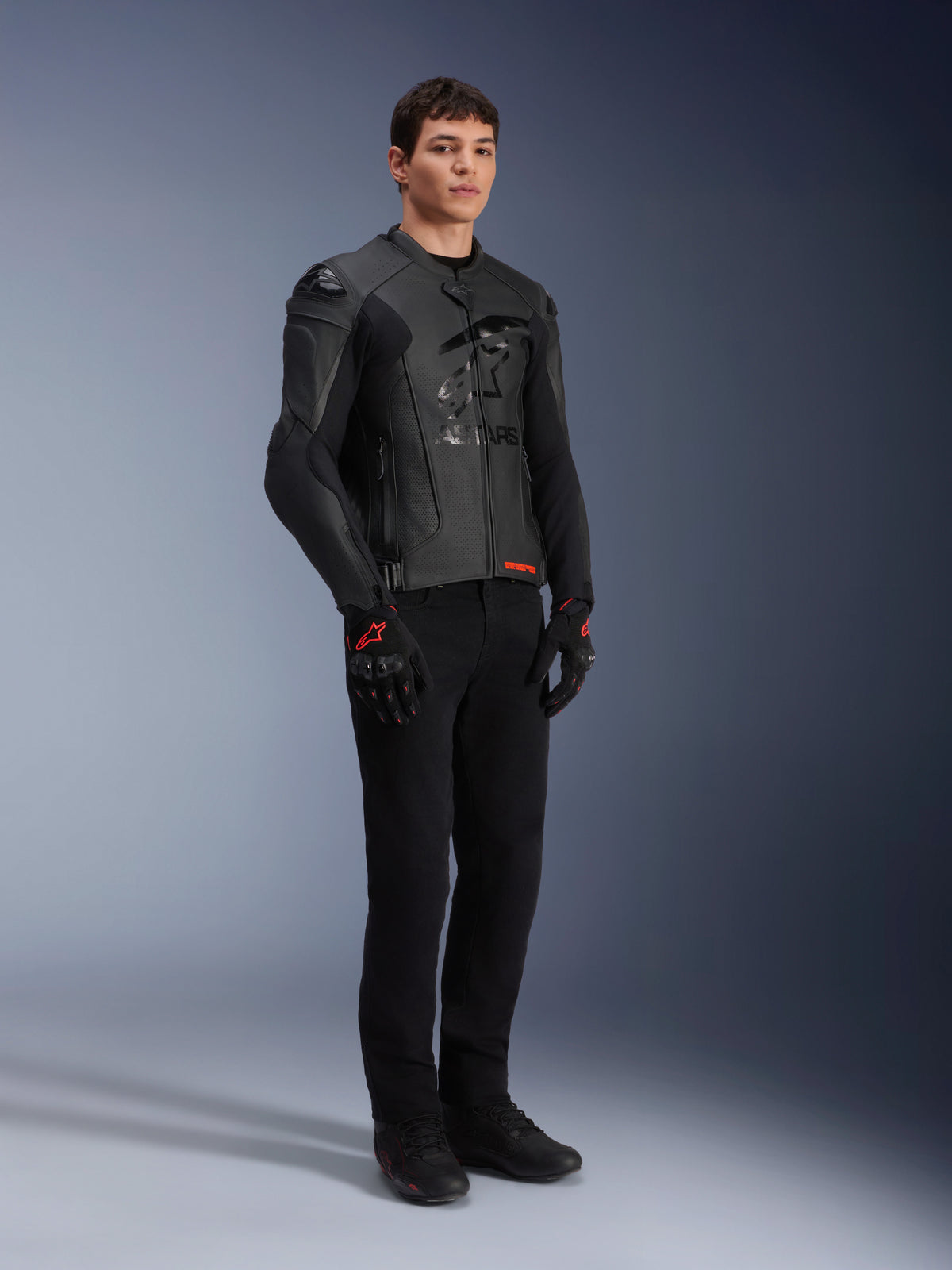 Casaco de couro Alpinestars GP Plus R V4 Airflow, preto, usado por um modelo masculino com calças de motociclismo pretas e luvas com detalhes vermelhos, com painéis de couro perfurado e sliders de ombro integrados, fotografia de estúdio de corpo inteiro.