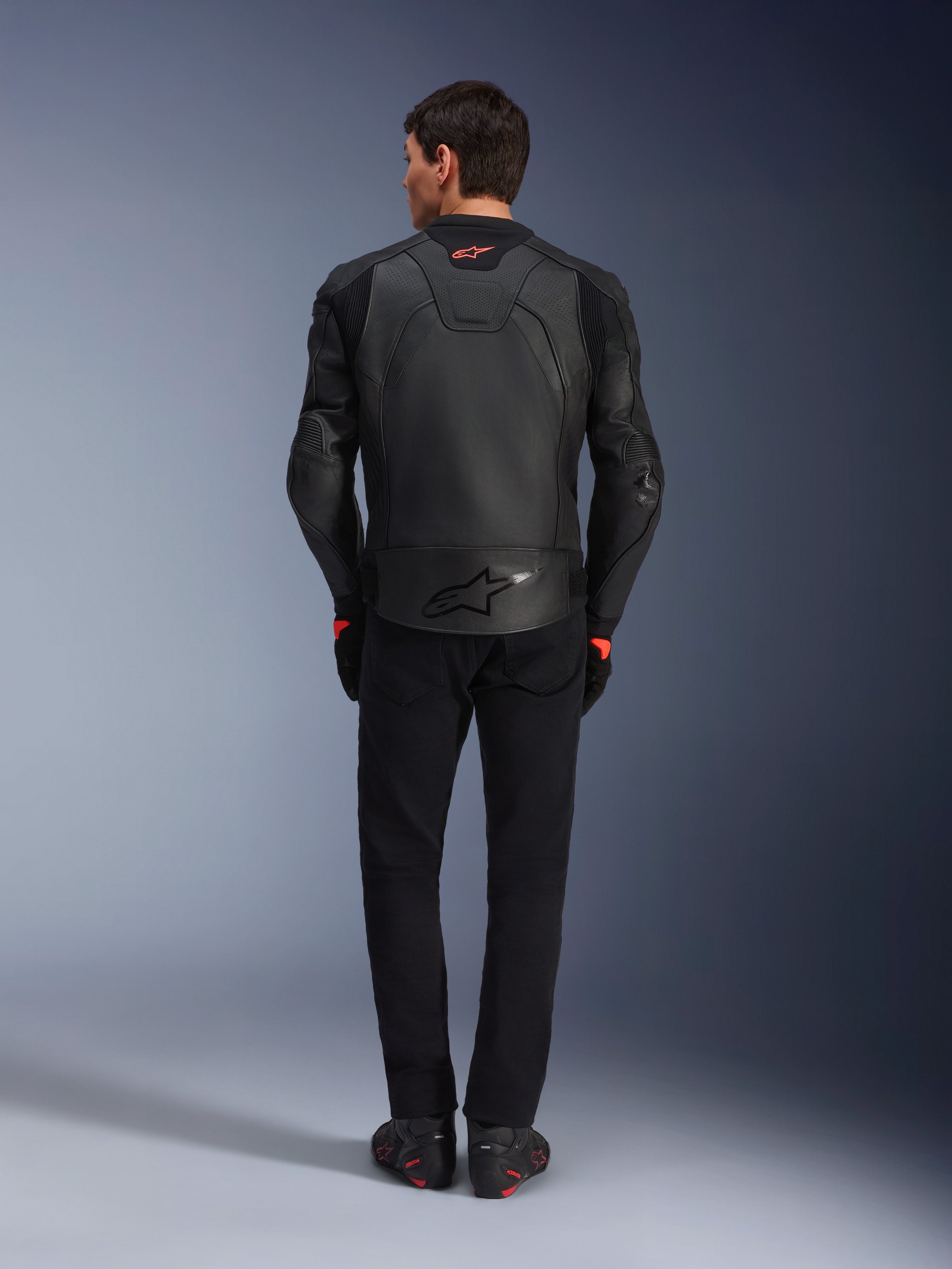 Jaqueta de Couro Alpinestars GP Plus R V4 Airflow, preta, vista traseira de piloto com equipamento completo, apresenta couro perfurado para ventilação e detalhes do logótipo em vermelho, design desportivo elegante, combinada com calças pretas e botas de motociclismo