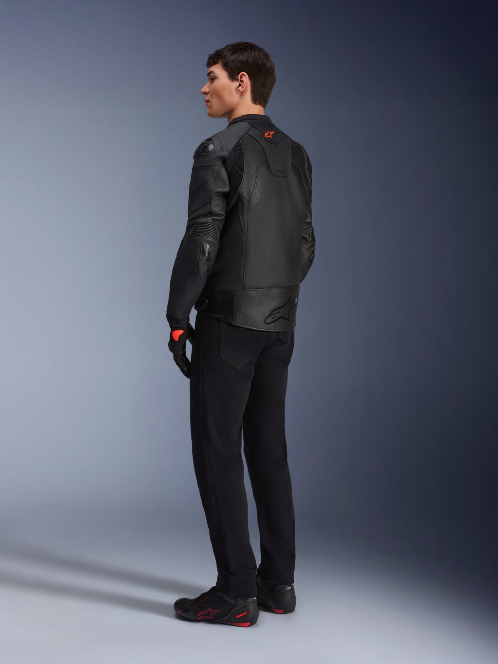 Jaqueta de Couro Alpinestars GP Plus R V4 Airflow, jaqueta de motociclismo desportiva para homem, totalmente preta, vista traseira de três quartos num modelo, com painéis de couro perfurado, logótipo vermelho nas costas e proteção de ombros integrada, usada com calças pretas e sapatos de condução