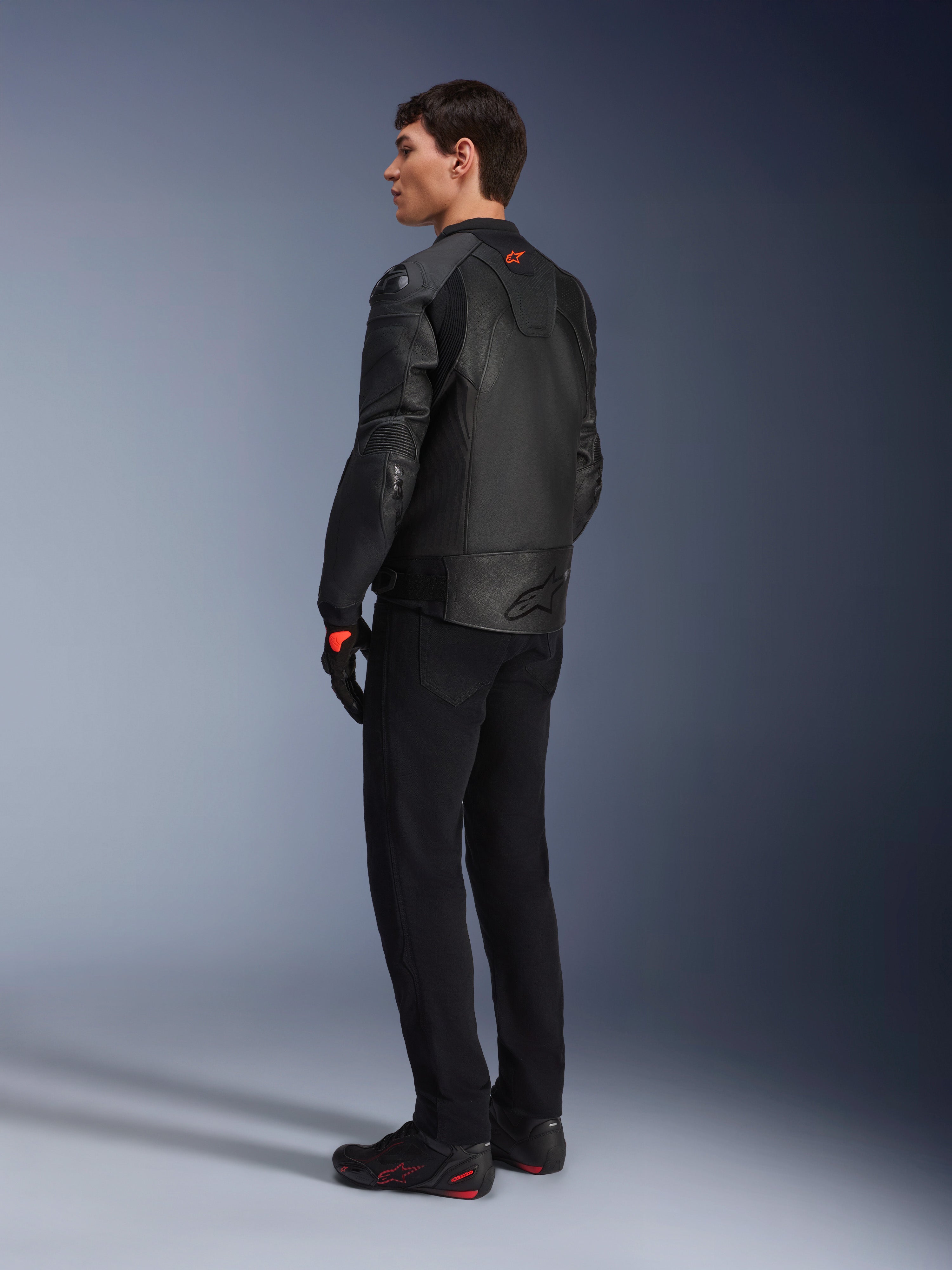 Jaqueta de Couro Alpinestars GP Plus R V4 Airflow, jaqueta de motociclismo desportiva para homem, totalmente preta, vista traseira de três quartos num modelo, com painéis de couro perfurado, logótipo vermelho nas costas e proteção de ombros integrada, usada com calças pretas e sapatos de condução