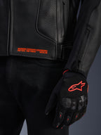 Alpinestars GP Plus R V4 Airflow Leather Jacket, grande plano de couro perfurado preto com texto gráfico cor-de-laranja, acompanhado por uma luva de motociclismo preta e cor-de-laranja com o logótipo Astars