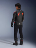 Rear view of a model wearing the Alpinestars GP Plus R V4 Airflow Leather Jacket, Black with Red Fluo and White accents, worn with black pants and black motorcycle boots, featuring prominent white logos on the back and sleeves, sport-style leather construction