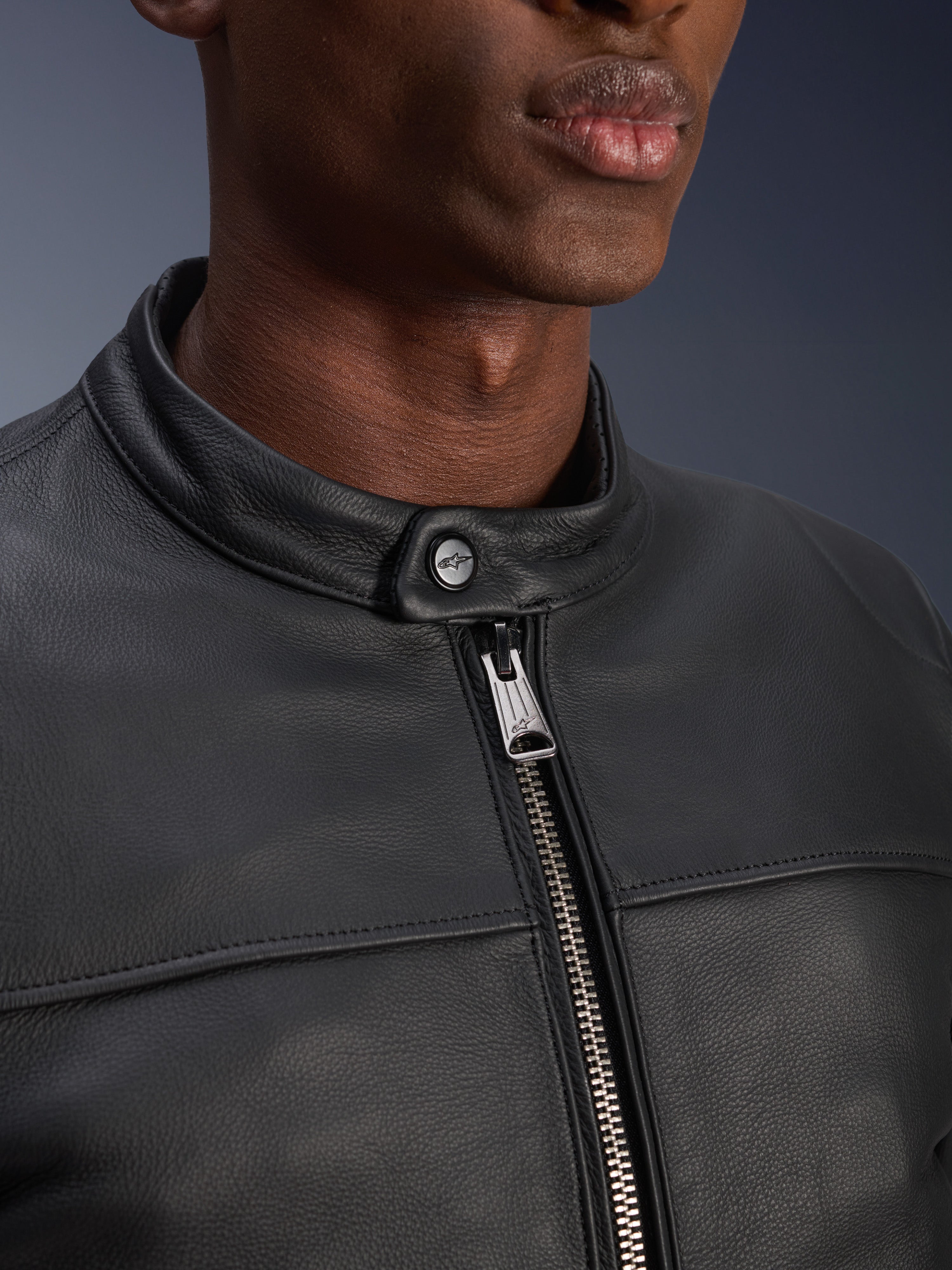 Close-up of the Alpinestars Blacktrack Leather Jacket, showing the metallic gunmetal zip and button neck closure with Alpinestars logo, premium black bovine leather texture, urban motorcycle apparel